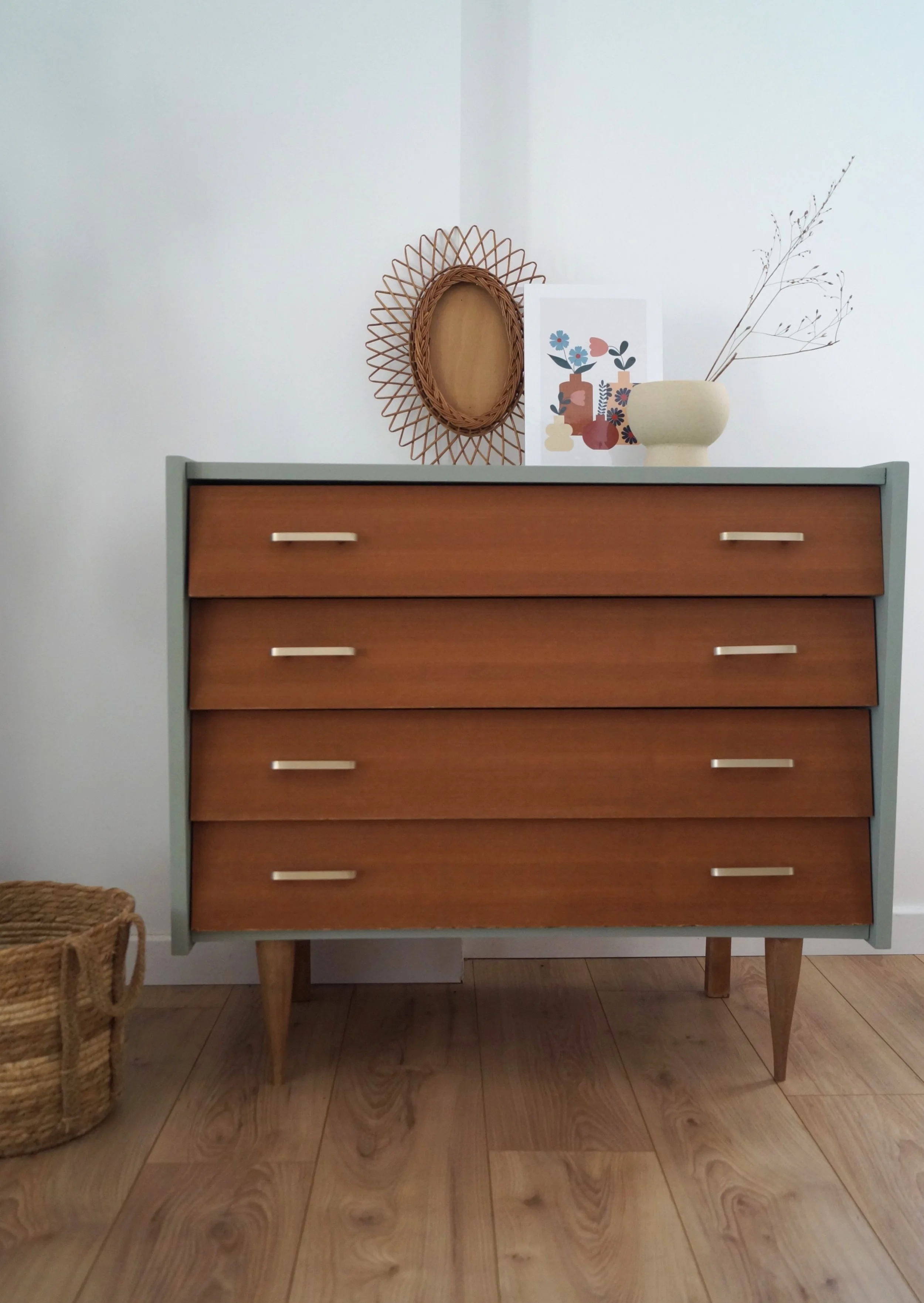 Une commode à quatre tiroirs en bois avec des poignées métalliques, sur un sol en bois clair, décorée avec un miroir en rotin, une affiche d'art abstrait, un vase beige avec des branches sèches et une panière en osier à gauche.