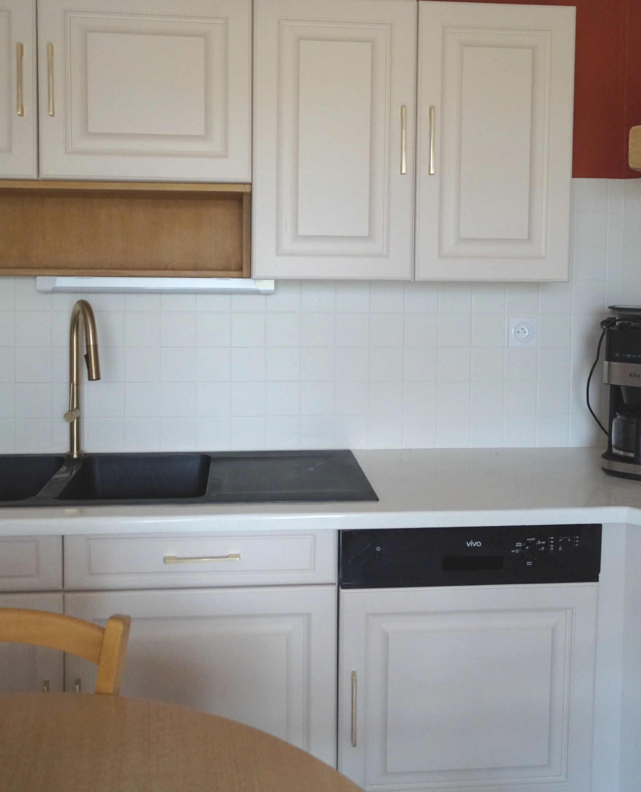 Cuisine blanche avec un évier en angle, un robinet en or, un lave-vaisselle noir, et une cafetière à droite.