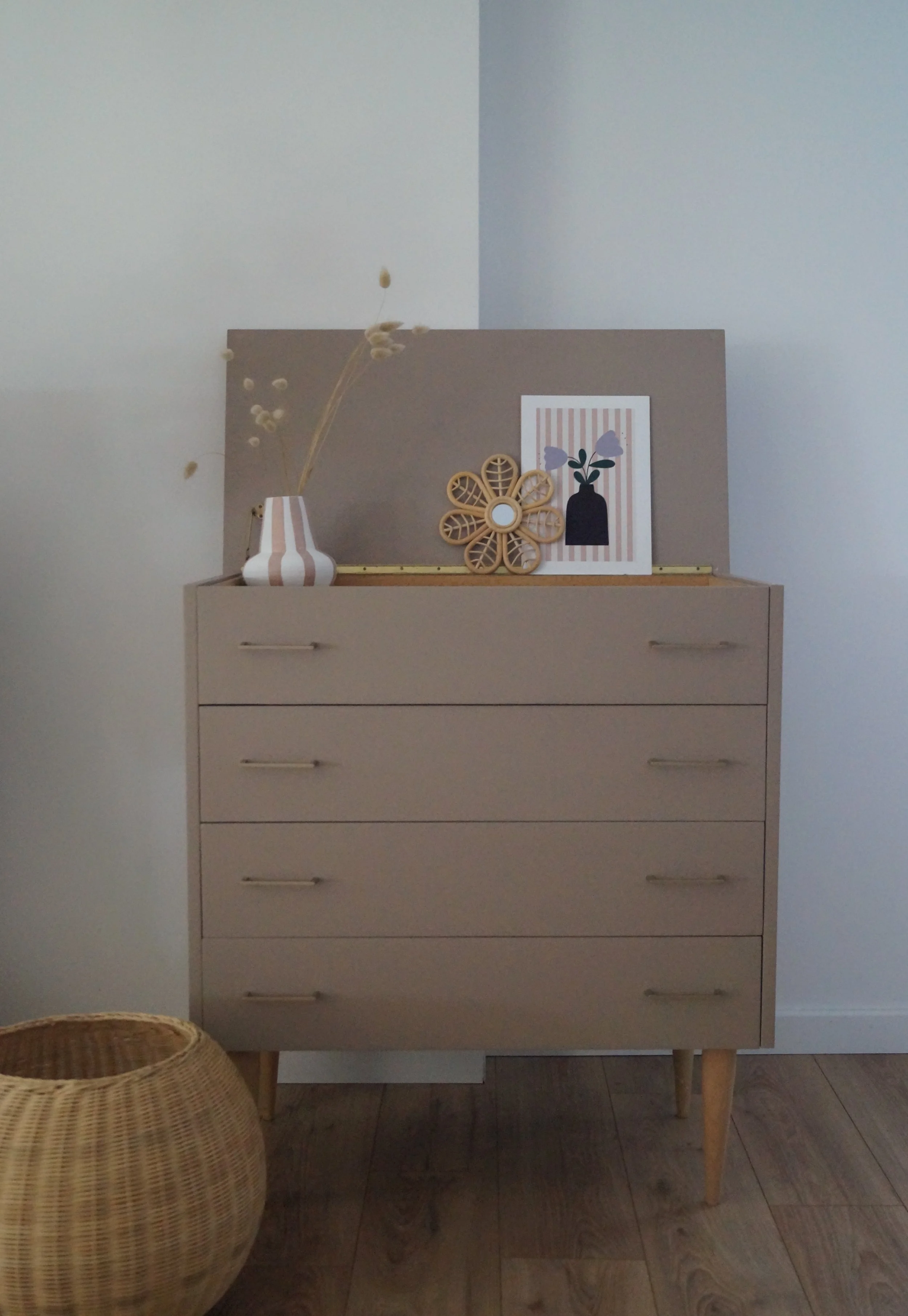 Une commode beige avec quatre tiroirs, décorée avec un vase blanc et beige, une décoration florale en bois, et une illustration d'une vase avec des fleurs, posés sur une surface avec un fond gris et blanc.