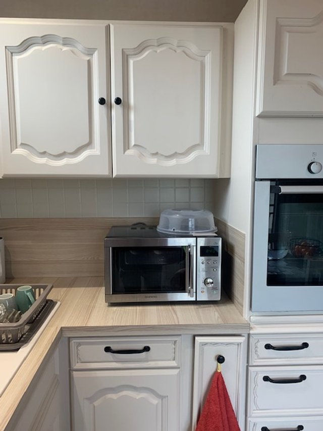cuisine avec des armoires blanches, un four électrique et un micro-ondes sur la counter, un torchon rouge accroché à la poignée d’un tiroir, et un plateau avec des tasses à café.