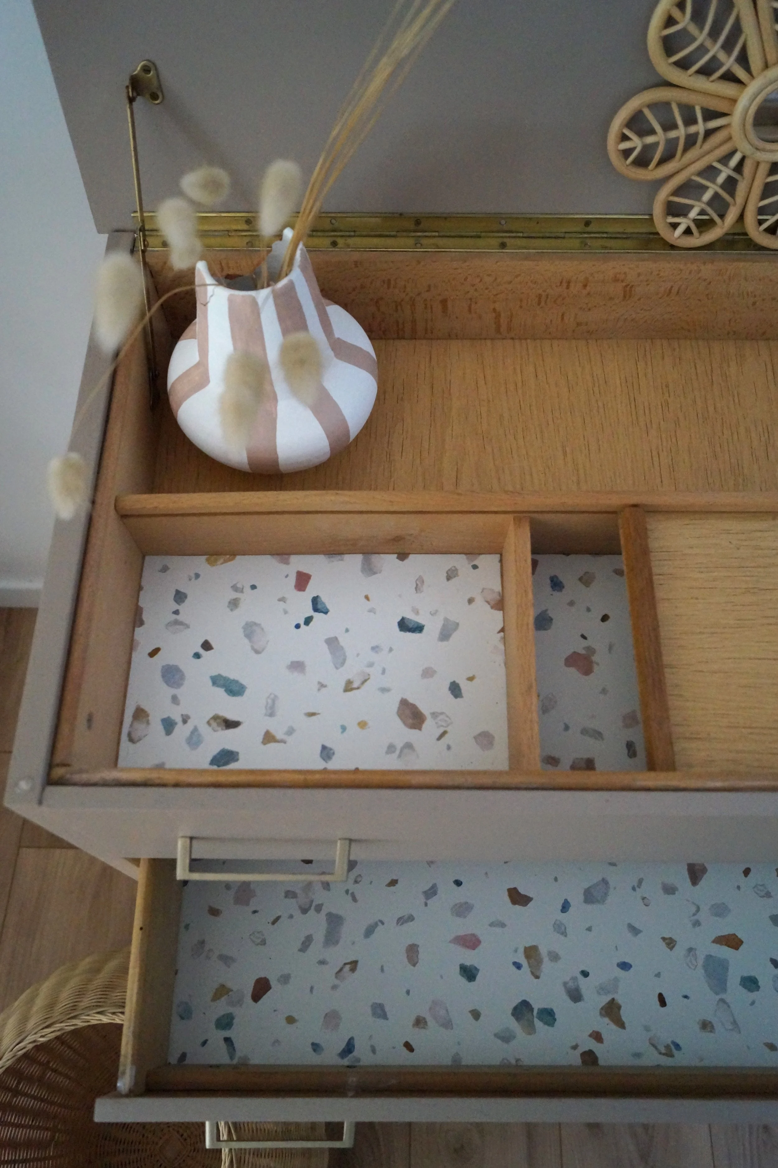 Vue d'une étagère en bois avec plusieurs compartiments, un vase blanc avec des rayures beiges contenant des tiges sèches et une décoration en forme de fleur en rotin.