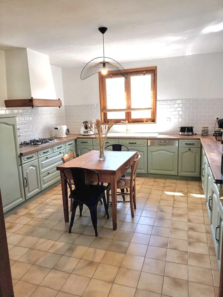 Cuisine avec armoires vertes, plan de travail en bois, motif de carreaux de sol en terre cuite, fenêtre en bois, table en bois avec un vase de fleurs séchées, quatre chaises, et appareils électroménagers.