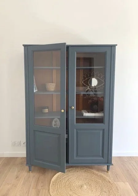 Vitrine bleue avec deux portes vitrées dans une pièce avec sol en bois et mur blanc, contenant des objets décoratifs