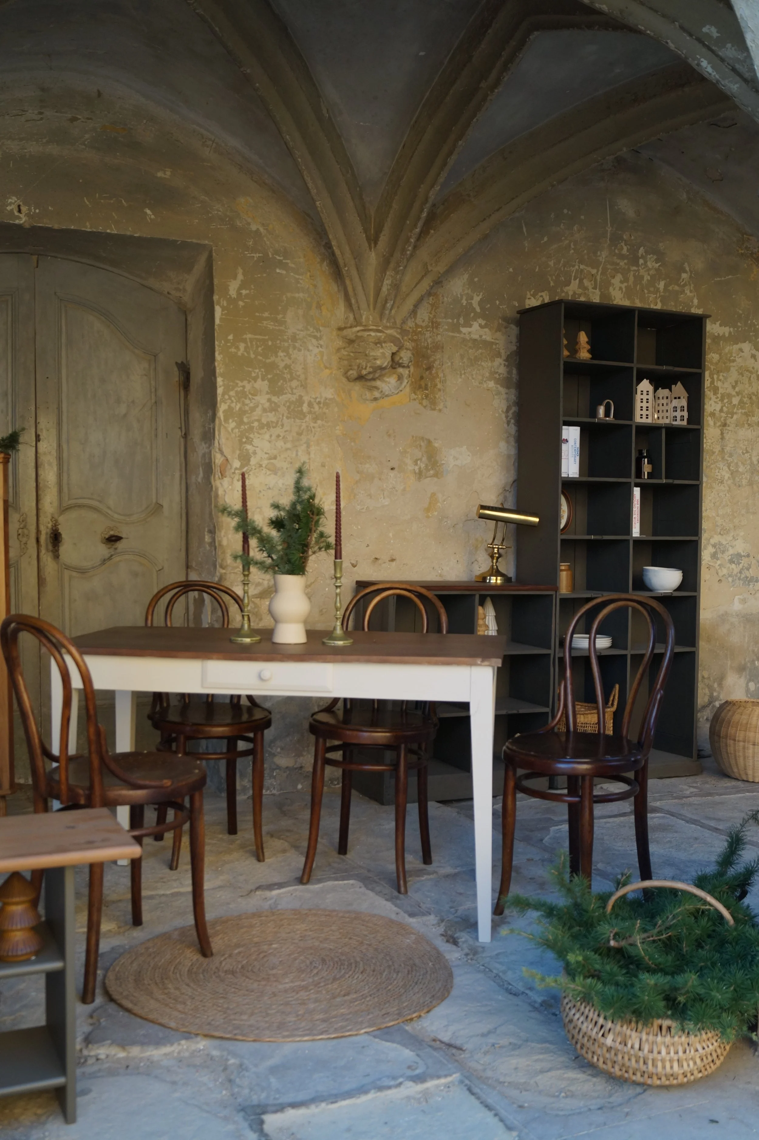Une pièce intérieure avec un mur en pierre vieilli, un plafond voûté et un vieux porte en bois. Il y a une table en bois blanc avec une vase contenant un petit sapin vert et deux chandeliers rouges. Autour de la table, il y a plusieurs chaises en bois foncé. Sur le côté droit, une grande étagère noire avec des objets de décoration et de la vaisselle. Sur le sol, il y a un tapis rond en fibres naturelles et un panier avec un petit sapin en decoration.