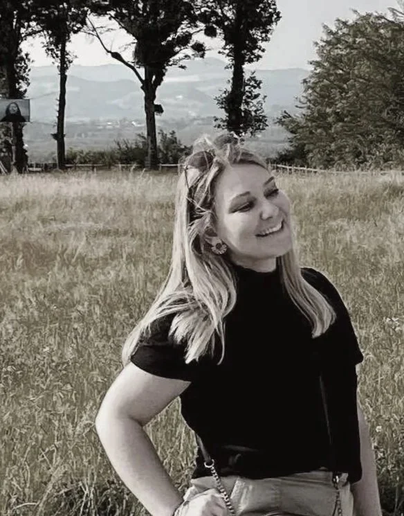Une jeune femme souriante avec des cheveux longs, portant un t-shirt noir, se tenant dans un champ herbeux avec des arbres et un paysage de montagnes en arrière-plan.
