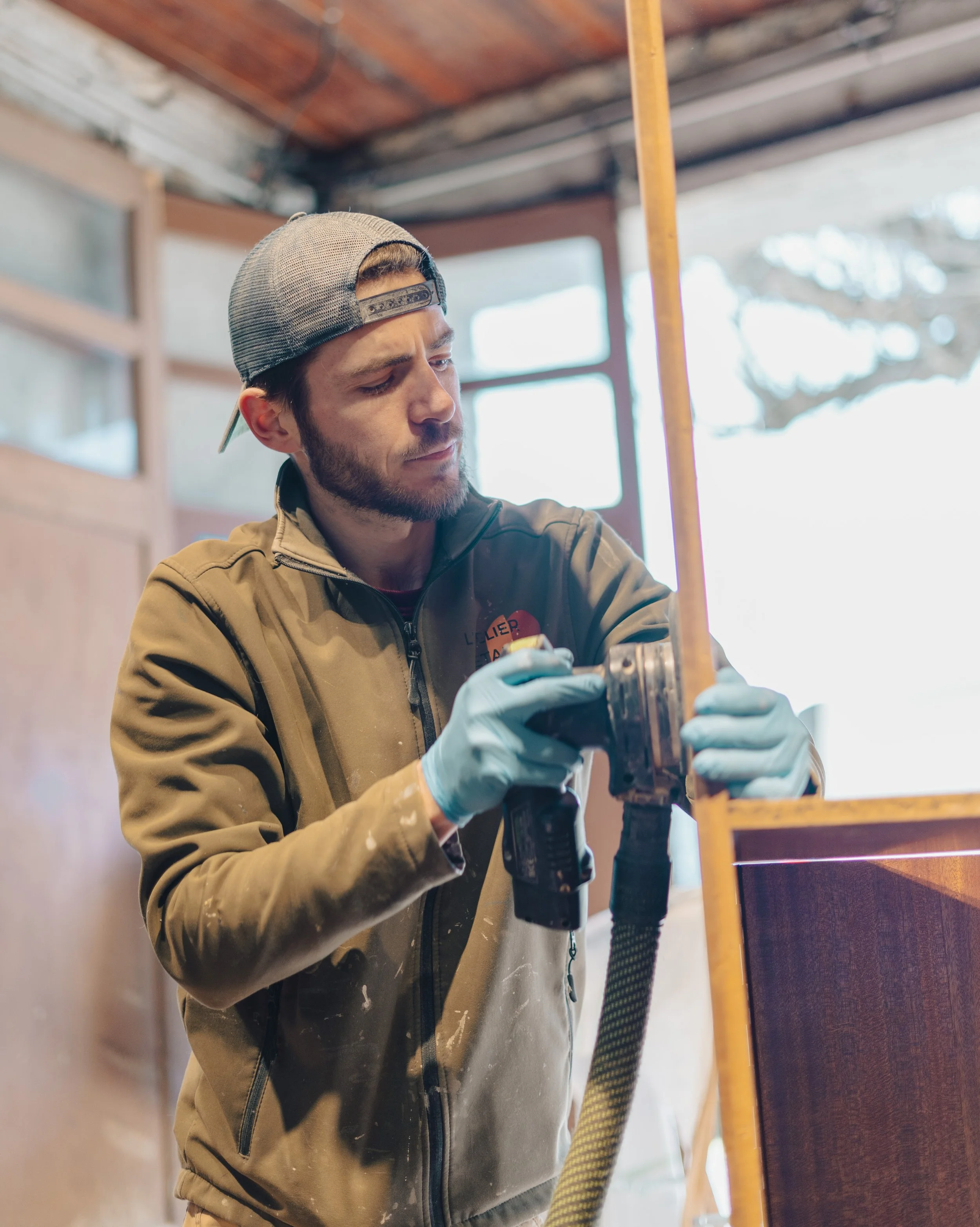 Un homme jeune portant un sweat beige, une casquette et des gants bleus, utilisant une perceuse électrique lors d'un travail de menuiserie ou de construction dans un atelier.