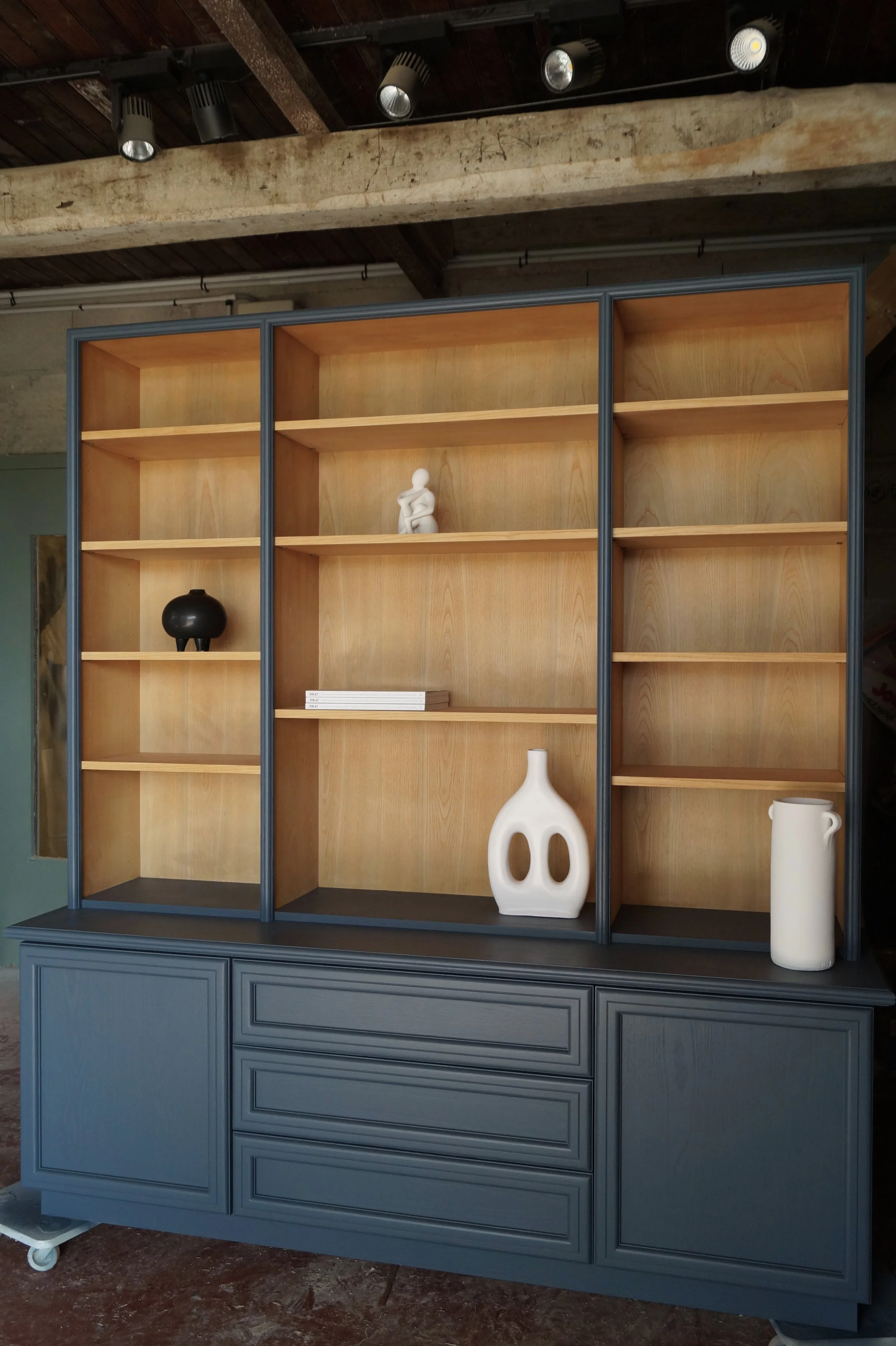 Une grande étagère en bois avec des portes bleues, contenant quelques objets décoratifs comme une petite sculpture blanche, une urne noire, une grande vase blanche et un autre vase blanc plus haut, dans une pièce avec des plafonds en bois et des spot