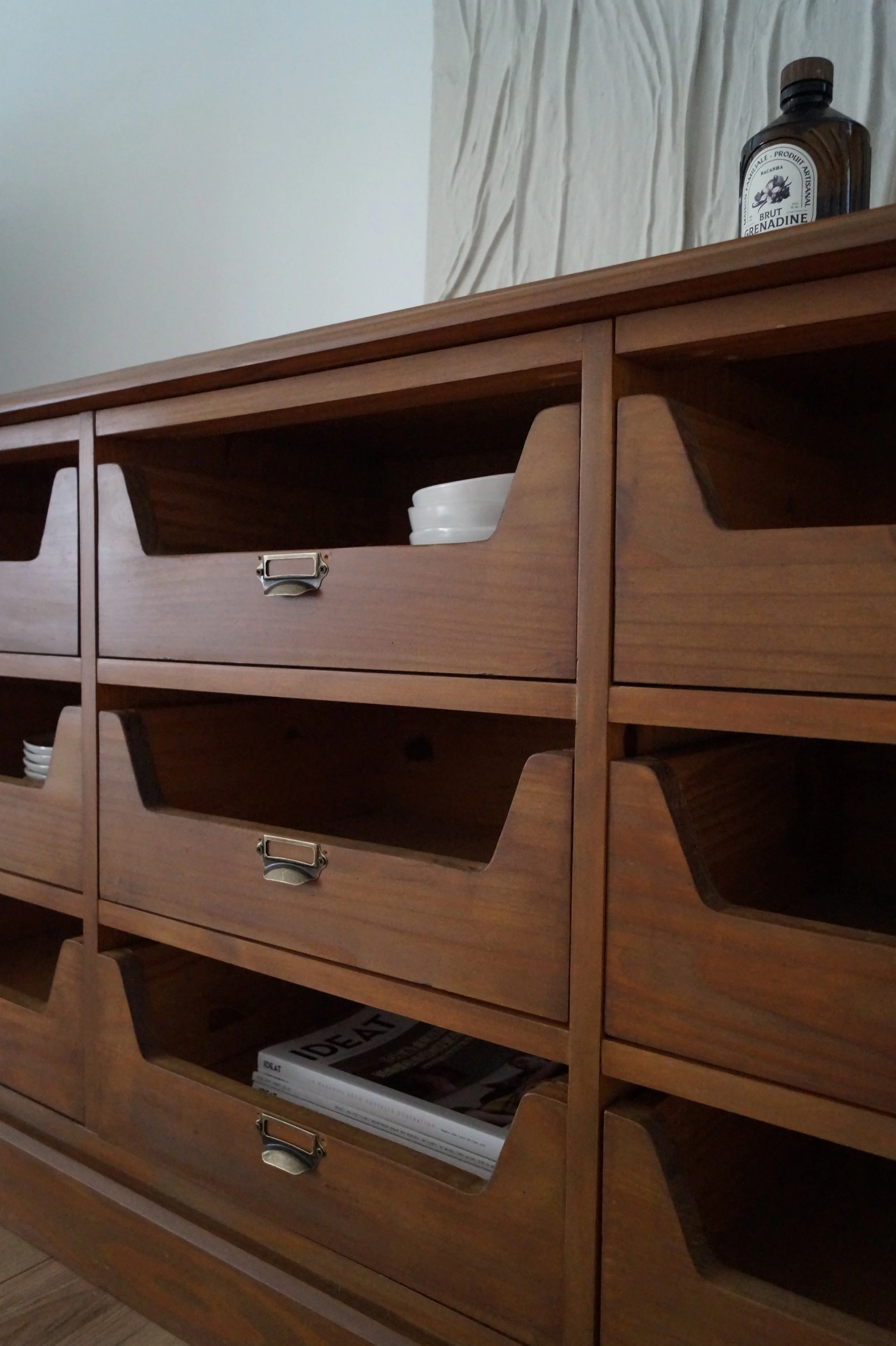 Une meuble en bois avec plusieurs tiroirs, certains tiroirs sont ouverts et contiennent des assiettes en plastique blanc. En haut du meuble, une bouteille en plastique foncée avec une étiquette