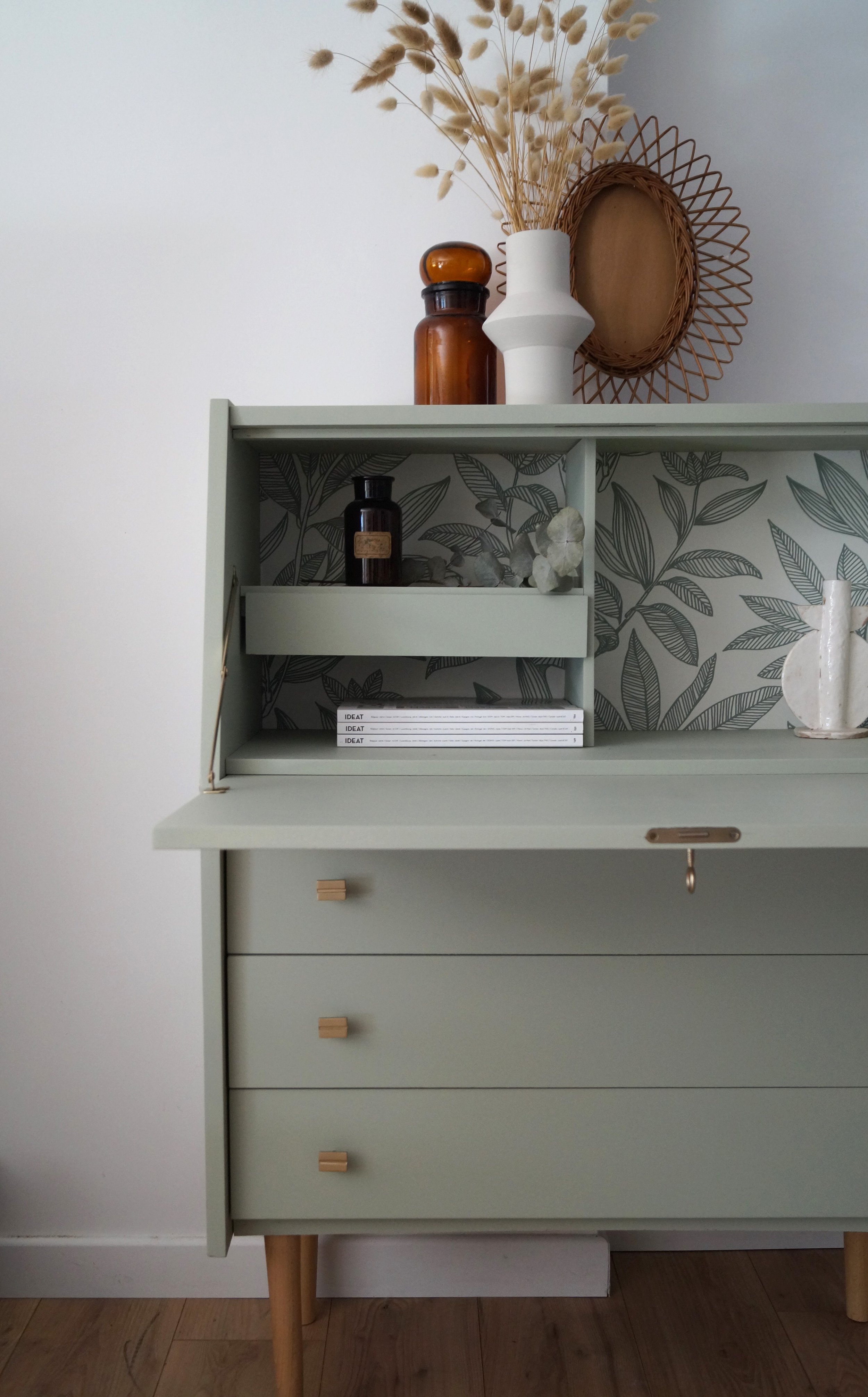 Un secrétaire vert pastel avec de petites poignées dorés, décorée de vases, de livres et d'objets en verre, avec un fond intérieur en motifs de feuilles.