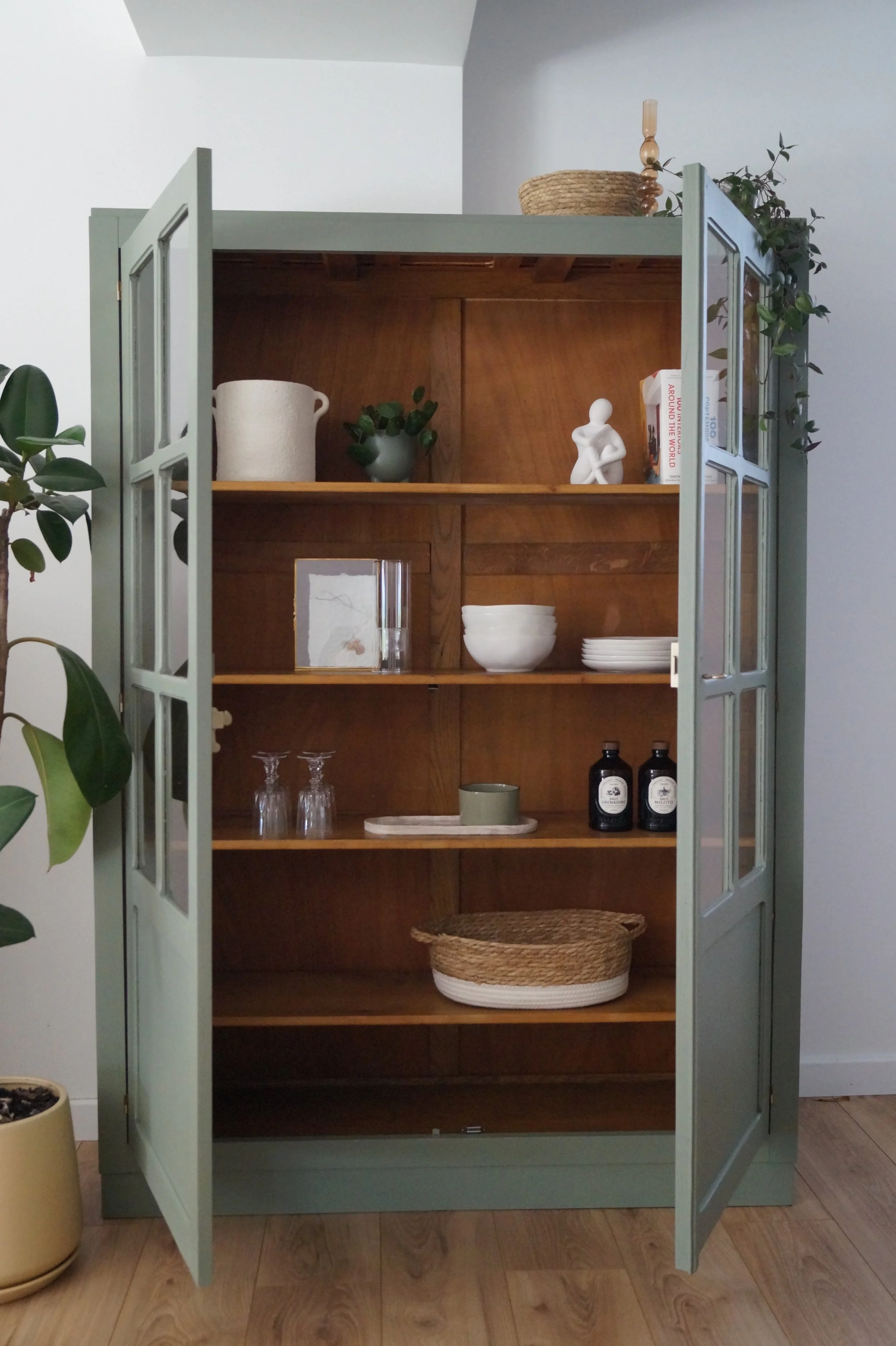 Une armoire en bois de style vintage, de couleur vert, avec des portes vitrées, contenant des objets de décoration et de la vaisselle, dans une pièce avec un sol en bois et une plante verte à gauche.