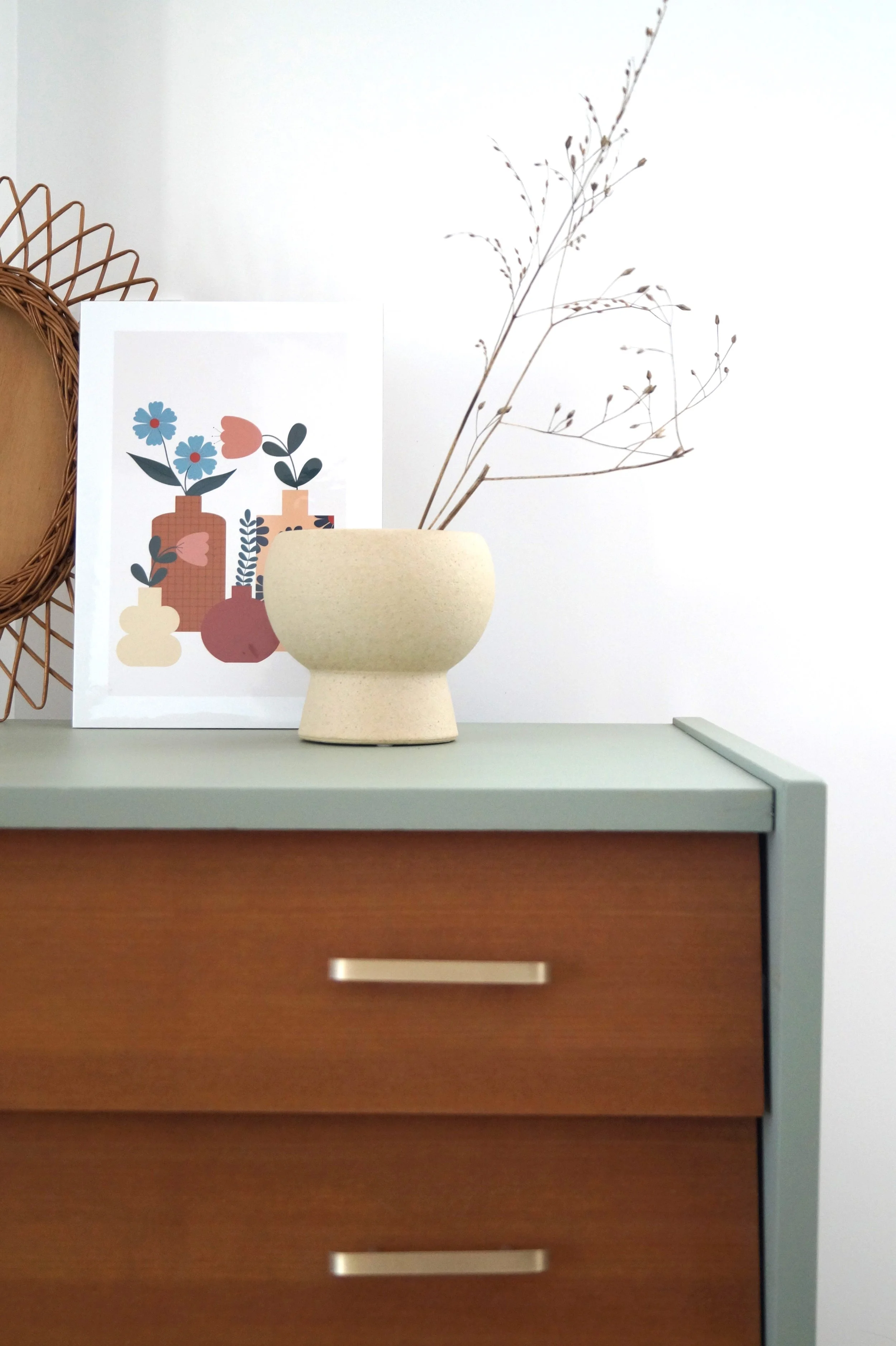 Une composition minimaliste sur une commode en bois avec un vase beige contenant des branches sèches, une affiche illustrée de vases colorés avec des fleurs, et un élément en rotin en arrière-plan.