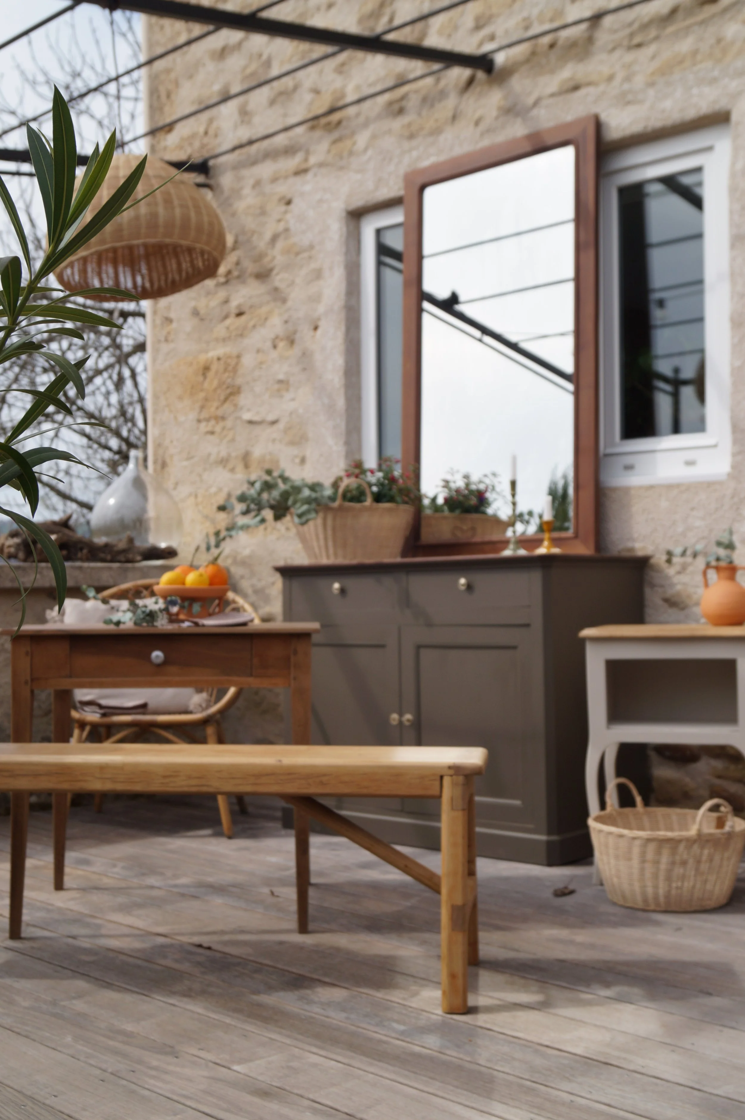Un balcon avec mobilier en bois, un miroir accroché au mur, une plante verte dans un pot, un buffet gris, une table avec fruits, un vase en terre cuite, un panier en osier, et une maison en pierre en arrière-plan.