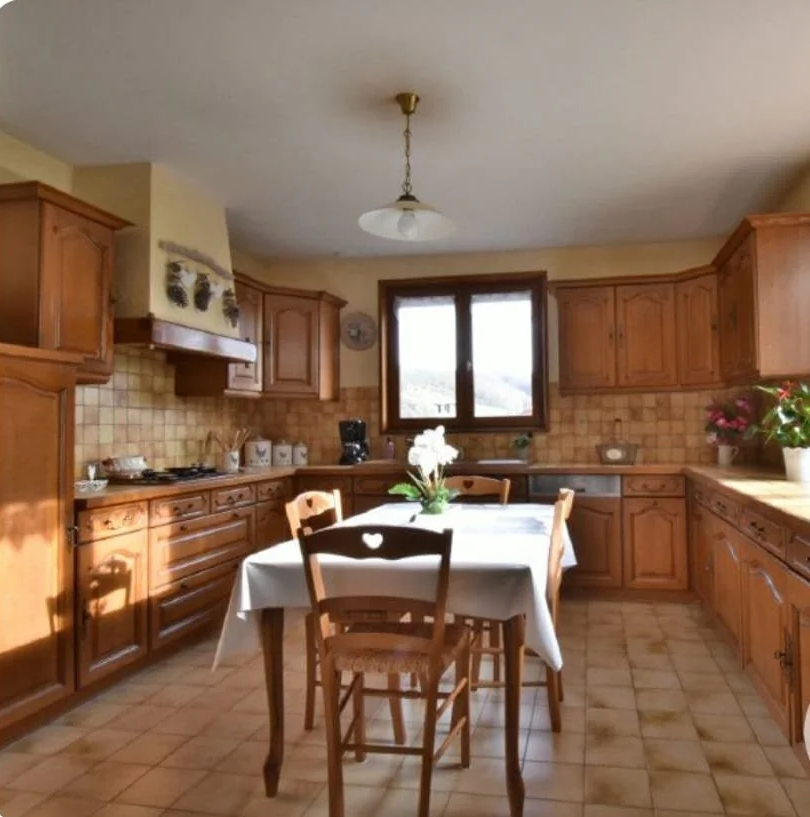Une cuisine rustique avec des meubles en bois, une table ronde avec une nappe blanche et des chaises en bois, une fenêtre derrière la table, des plantes en pot, un luminaire suspendu au plafond, et un plan de travail en bois.