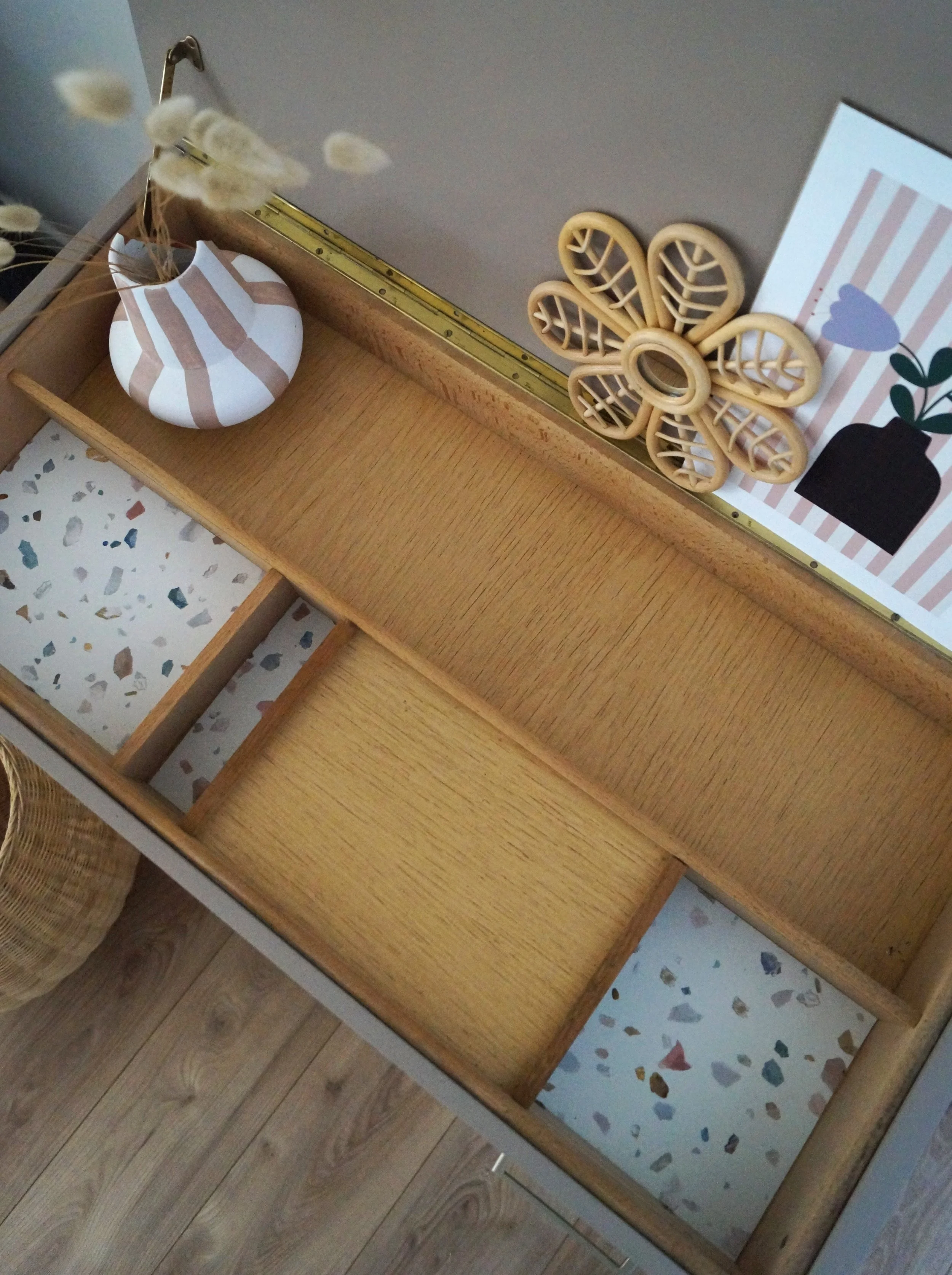 Un tiroir en bois avec plusieurs compartiments, décoration avec une figure de vases en céramique, une plante en papier décorative, un tableau avec une pot de fleurs en illustration, et une règle de métal