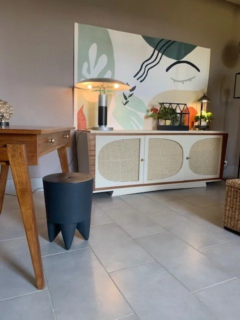 Intérieur contemporain avec un meuble de rangement blanc, une large peinture abstraite aux formes organiques, une lampe sur une table en bois, et un fauteuil en rotin sur un sol en carreaux gris.