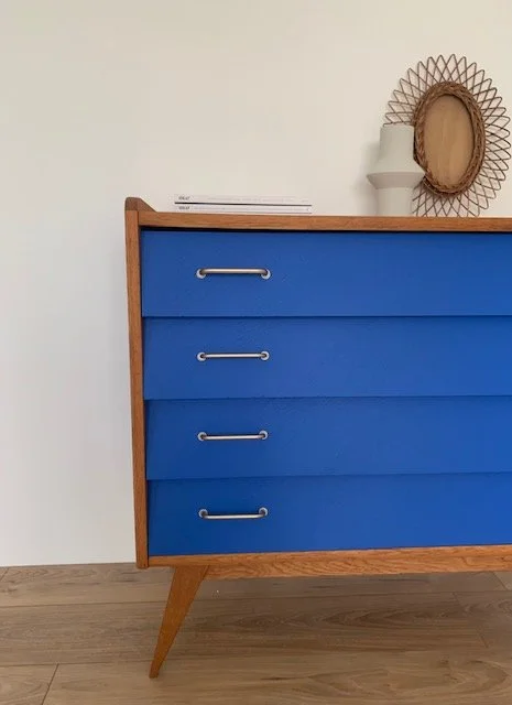 Commode bleue avec quatre tiroirs, décorée avec un miroir rond, un vase blanc et un livre, sur un fond blanc et un sol en bois.