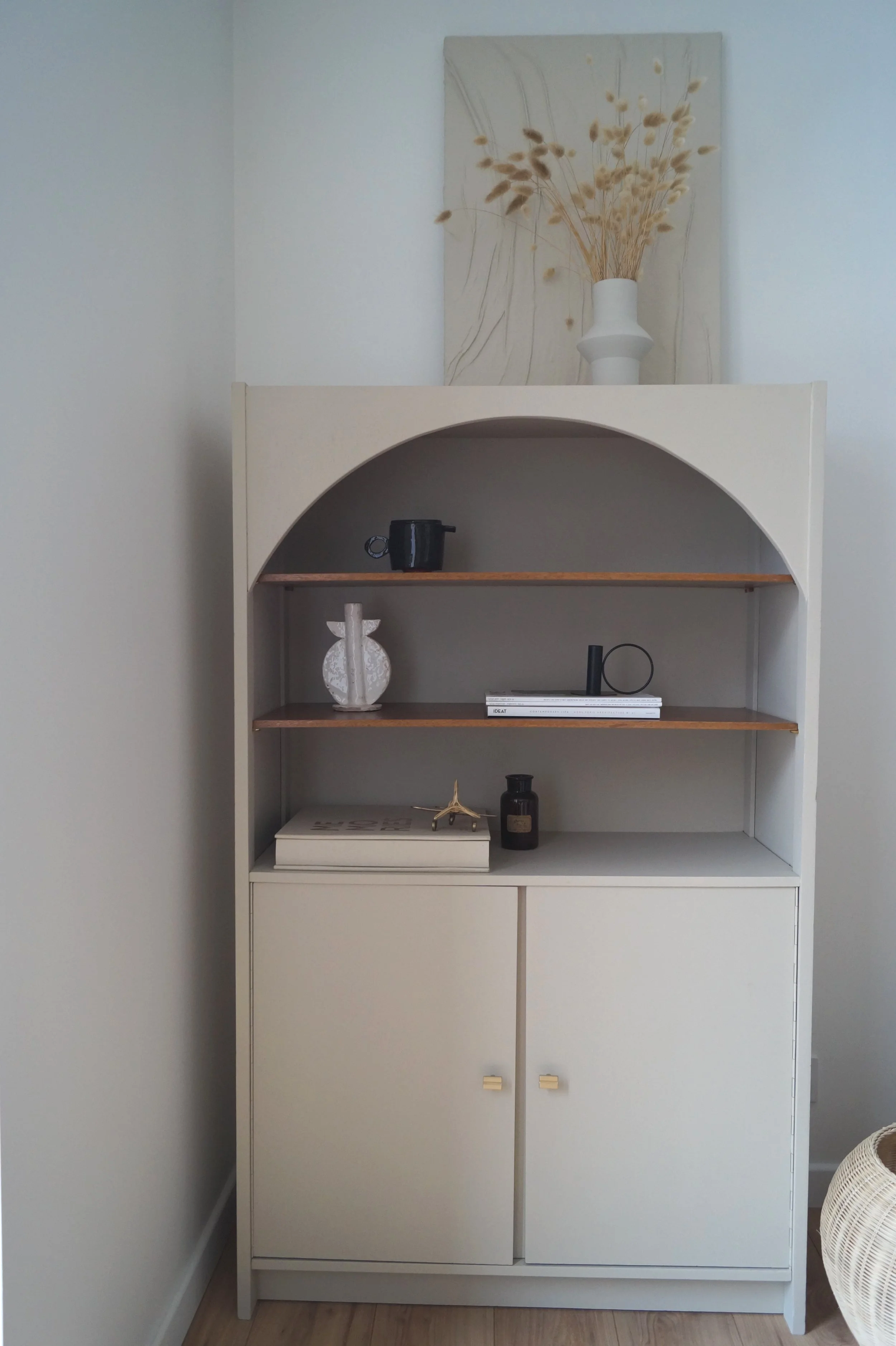 Une étagère blanche avec deux portes en bas, décorée avec des livres, des vases et une petite figurine en forme d'avion, avec une peinture de fleurs séchées en arrière-plan.