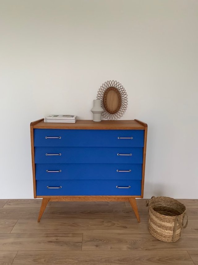 Commode en bois avec tiroirs bleus, une lampe blanche, un miroir tressé au-dessus, un livre, un vase blanc, un panier en osier à côté, sur un sol en bois.