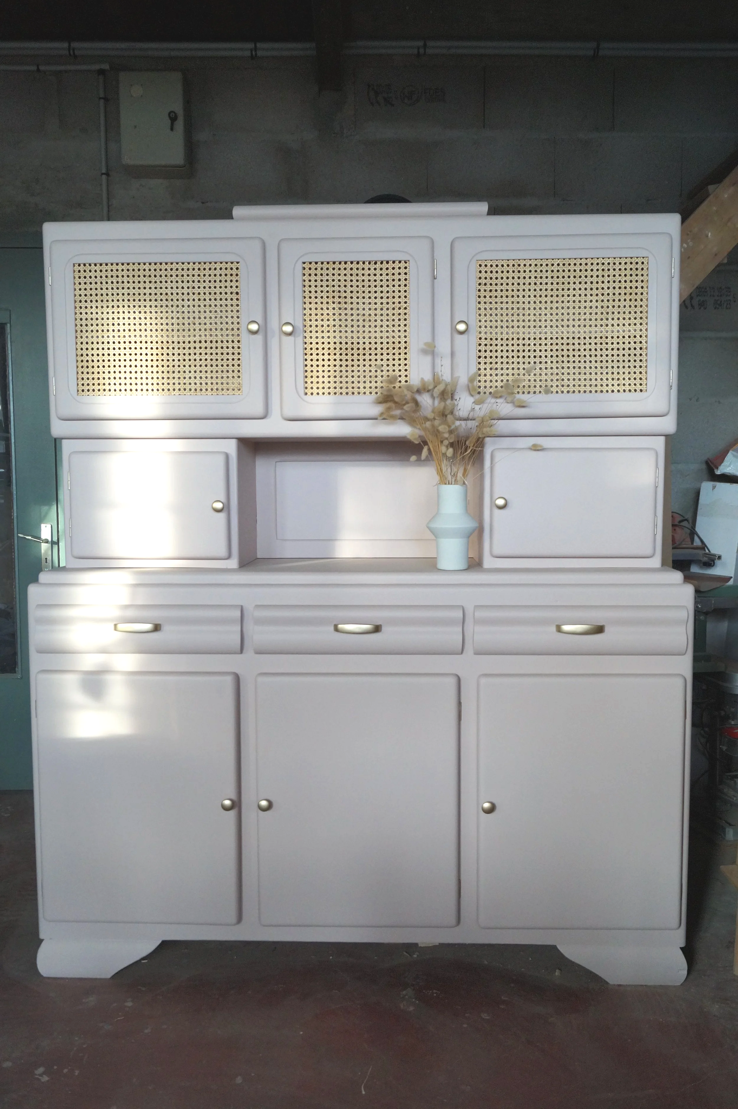 Un meuble rose pale ancien avec plusieurs portes et tiroirs, une vase blanche avec des fleurs séchées dessus.