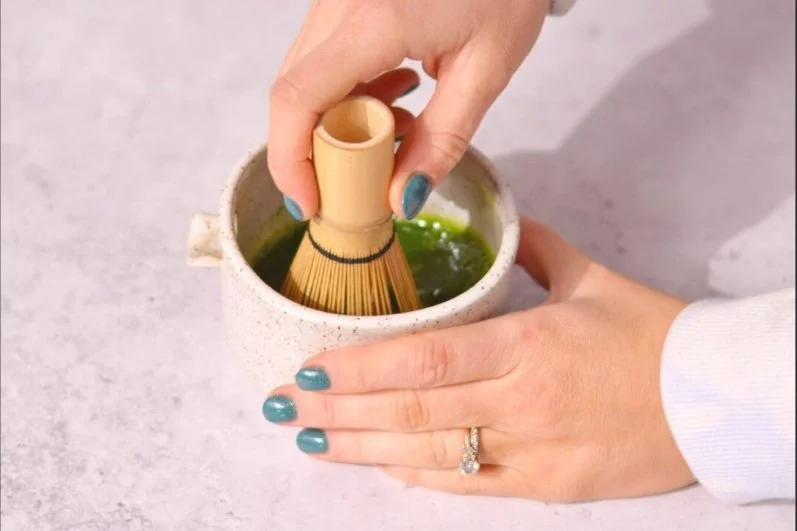 Person holds bamboo whisk and stirs matcha green tea in ceramic cup
