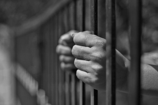 A person in a prison cell. A closeup of hands holding prison bars