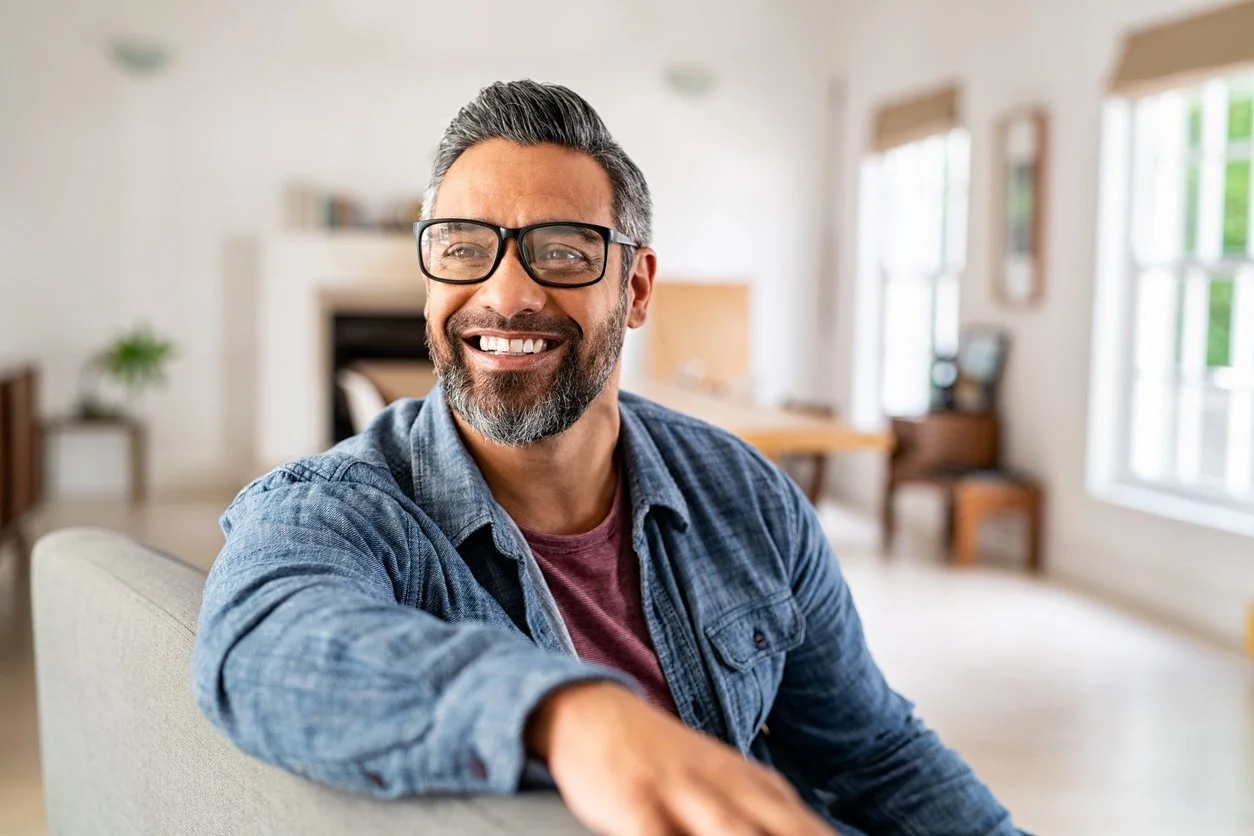 A man, wearing glasses and a blue button down shirt, smiling while sitting on a couch in a living room