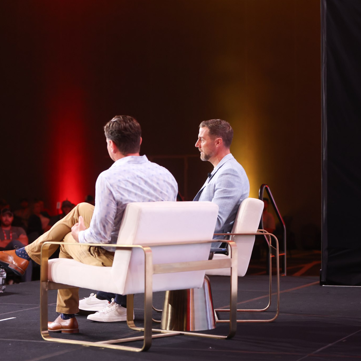 Two men seated on a stage in conversation during a conference or panel discussion, with an audience visible in the background.