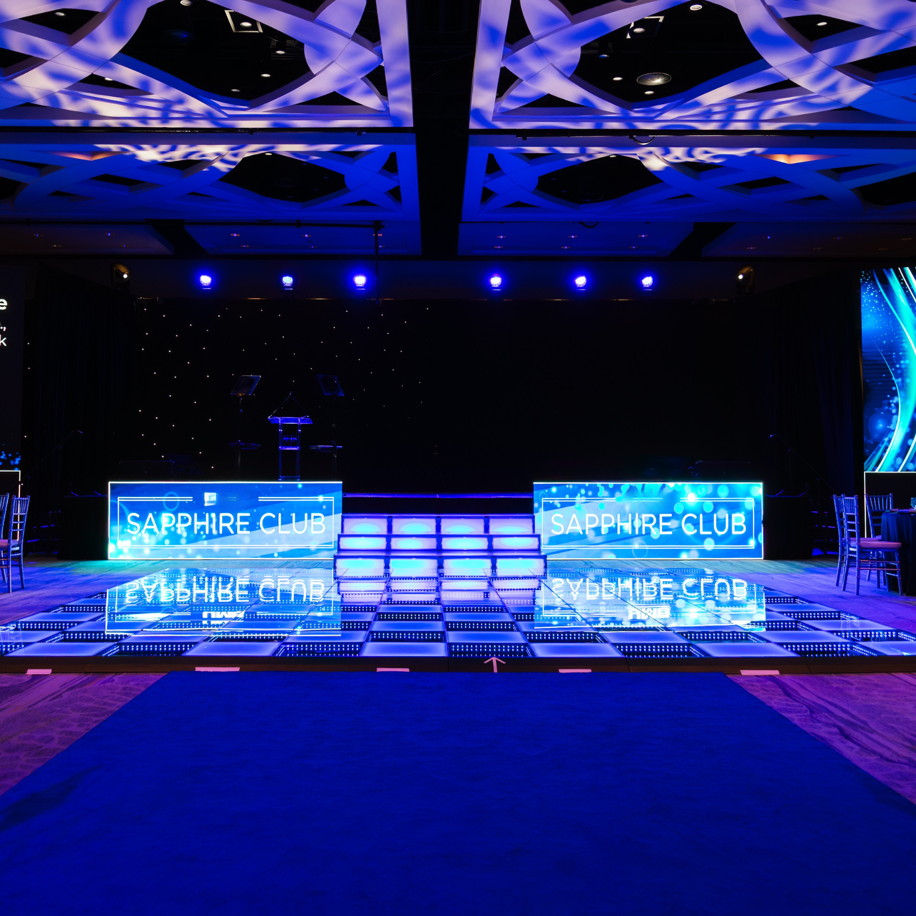 Scenic landscape featuring a large body of turquoise water in the foreground, rolling hills with grass and sparse bushes, and mountains with rocky peaks under a cloudy sky in the background.