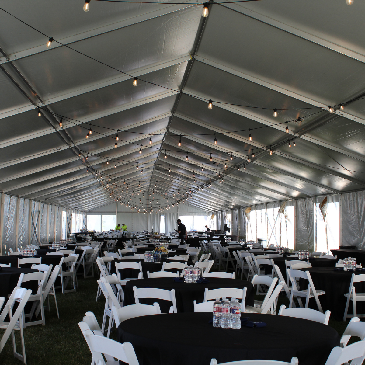 Inside a large event tent with string lights hanging from the ceiling, arranged tables with black tablecloths, white chairs, and bottles of water on each table, ready for a gathering or celebration.