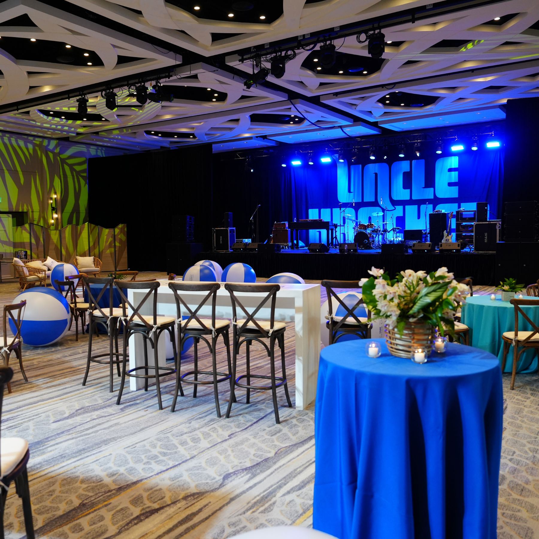 An event space with a stage, musical instruments, and seating arrangement with tables, chairs, and balloons. Decor includes a blue tablecloth with a floral centerpiece and candles.