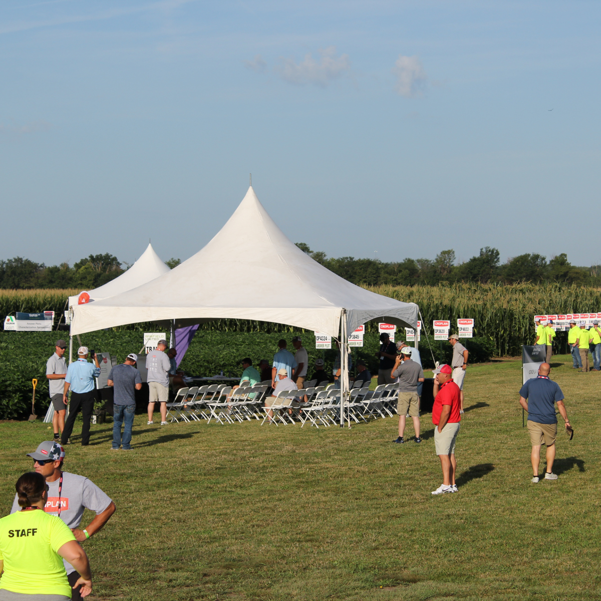 People gathered under a large white tent at an outdoor event in a field, with chairs arranged inside the tent and signs for crop varieties in the background.