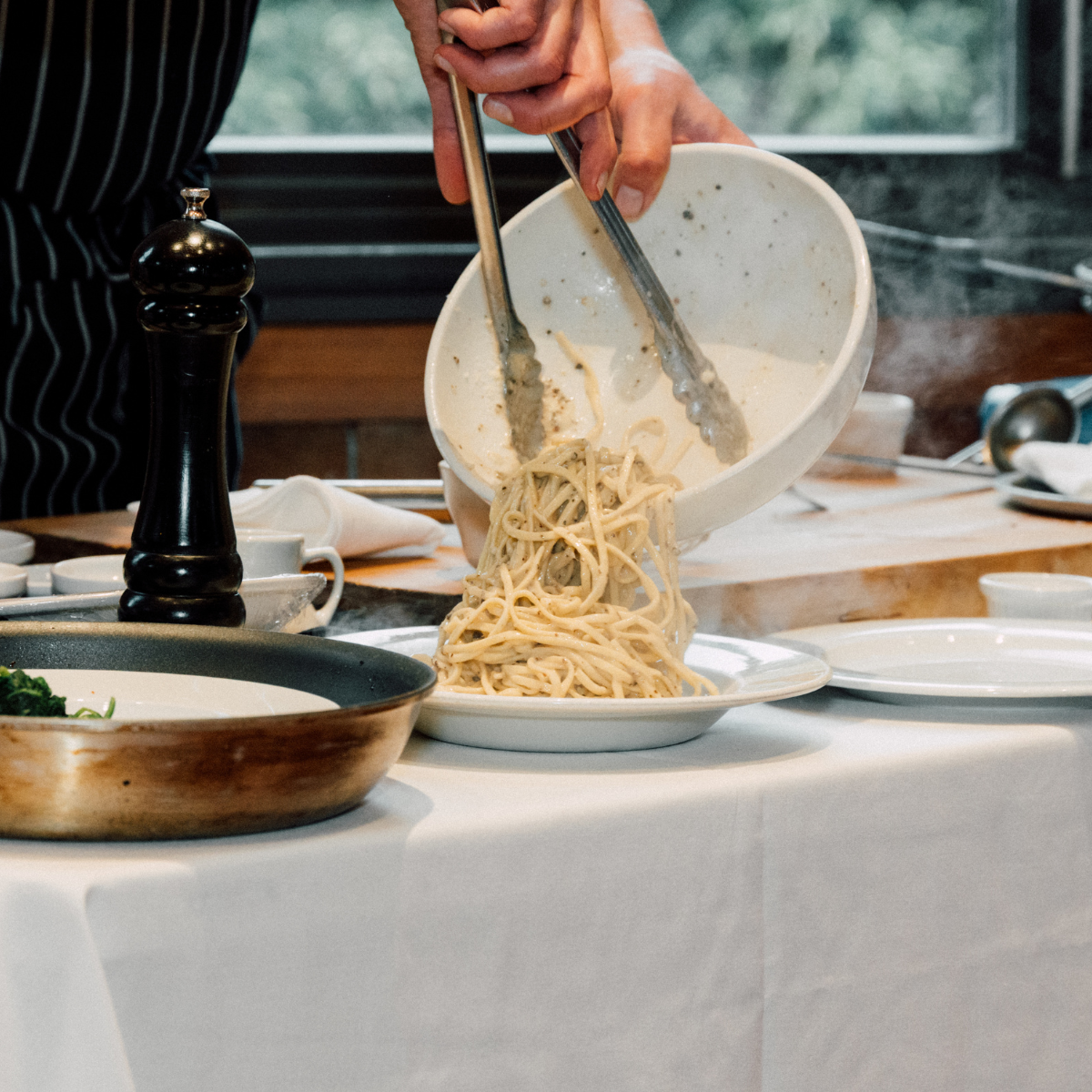 Person is serving pasta from a white bowl onto a plate, with steam rising in the background.