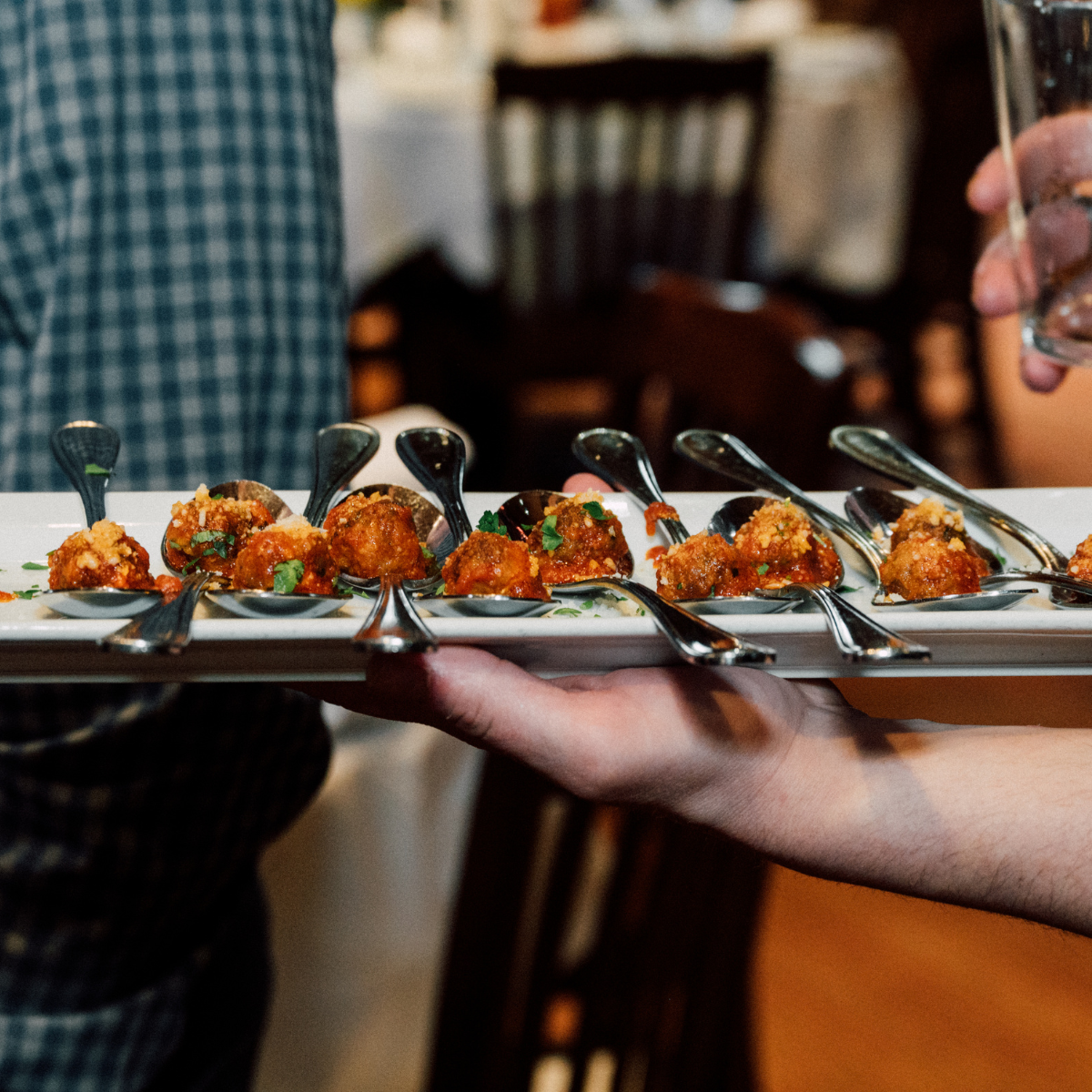 A server holding a white rectangular tray with several spoons, each with a portion of fried appetizer garnished with herbs.