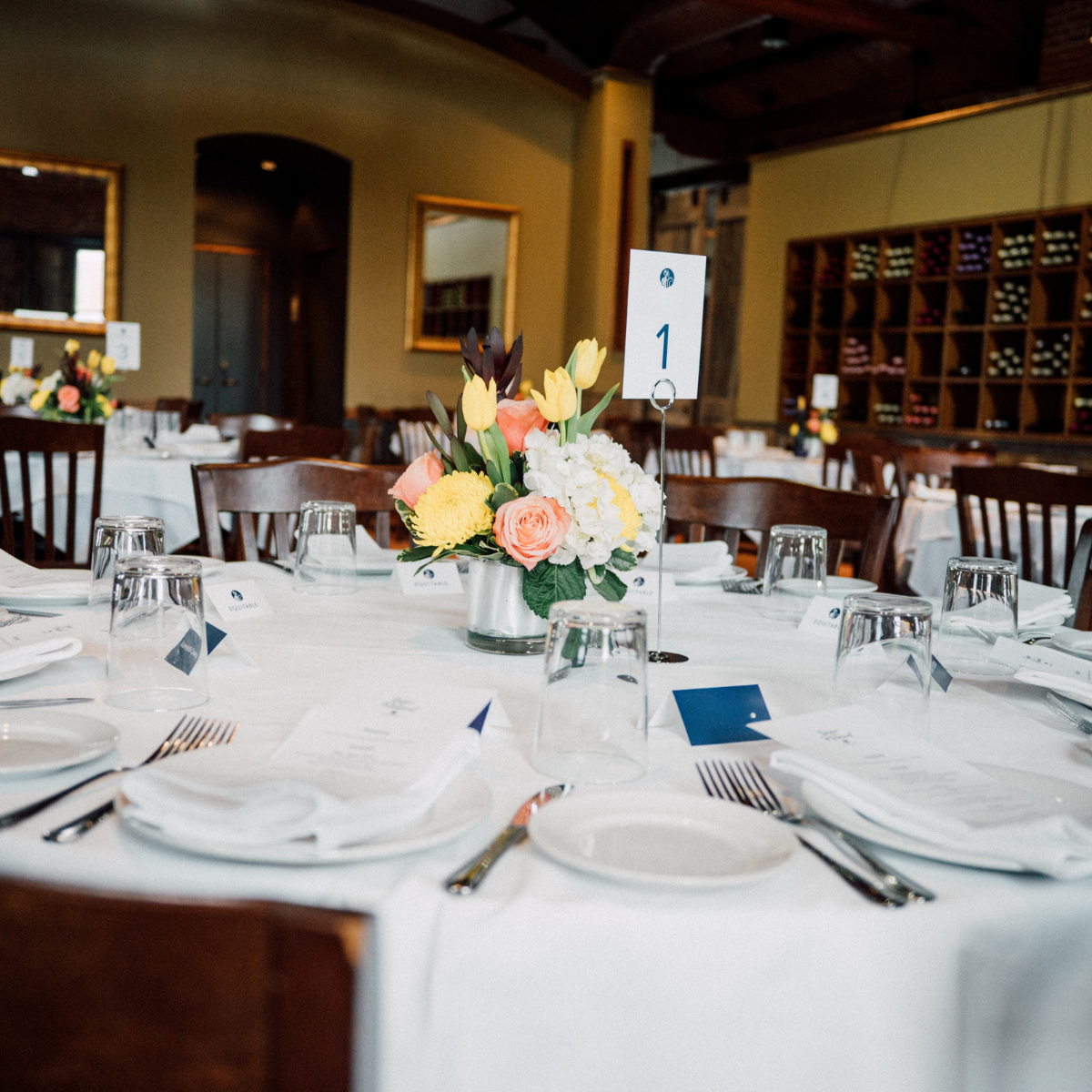 Wedding reception table with a centerpiece of yellow, peach, and purple flowers, set with white plates, silverware, and upside-down glasses. There is a table number sign showing number 1.