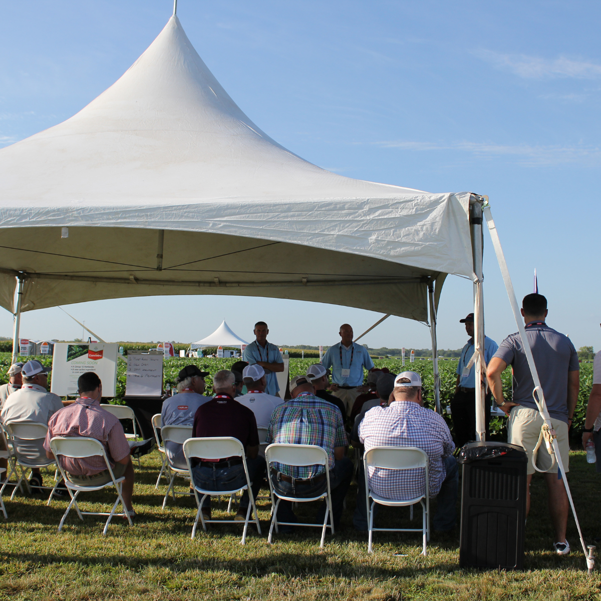 A group of people attending an outdoor presentation under a white canopy tent in a field, with speakers at the front and the audience seated on white folding chairs.