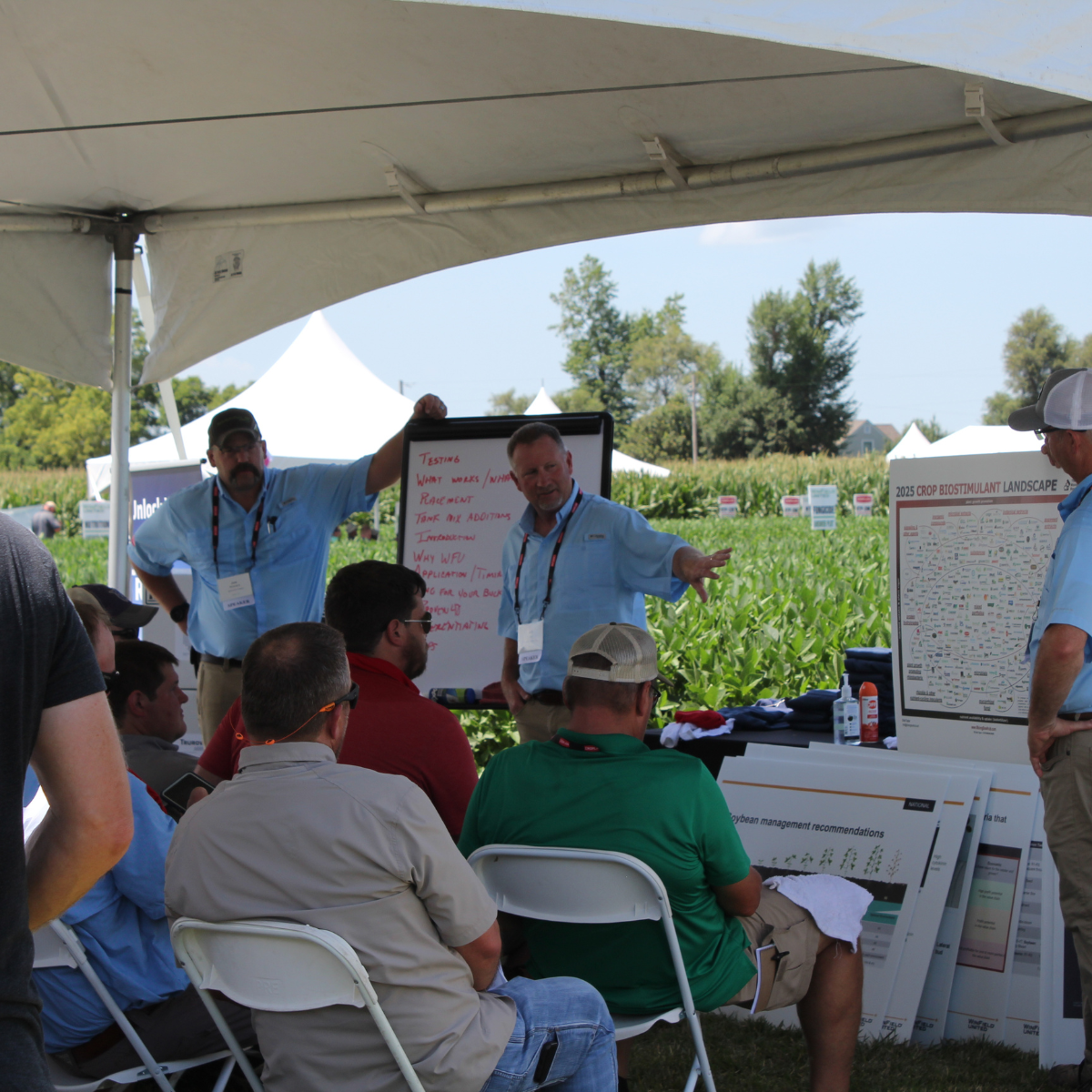 Group of people attending a presentation under a white tent outdoors, with farmland and crops in the background. Two men are standing near a whiteboard and a posters, speaking and gesturing to the audience.