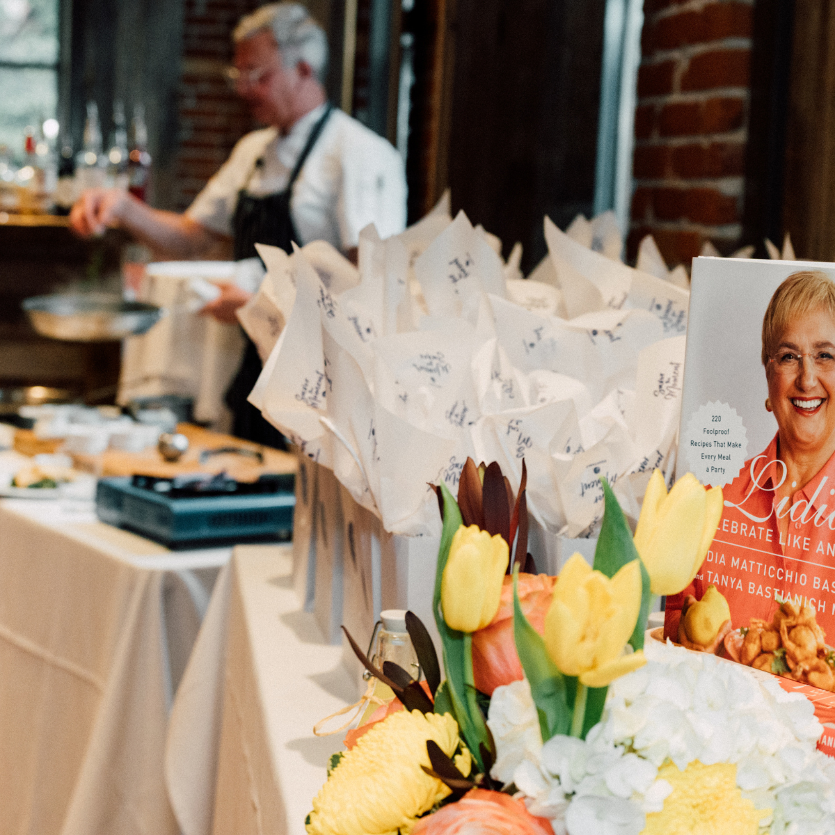 A blurred chef preparing food in a rustic kitchen with a table of wrapped food items in the foreground, decorated with colorful flowers and a promotional sign featuring a smiling woman.