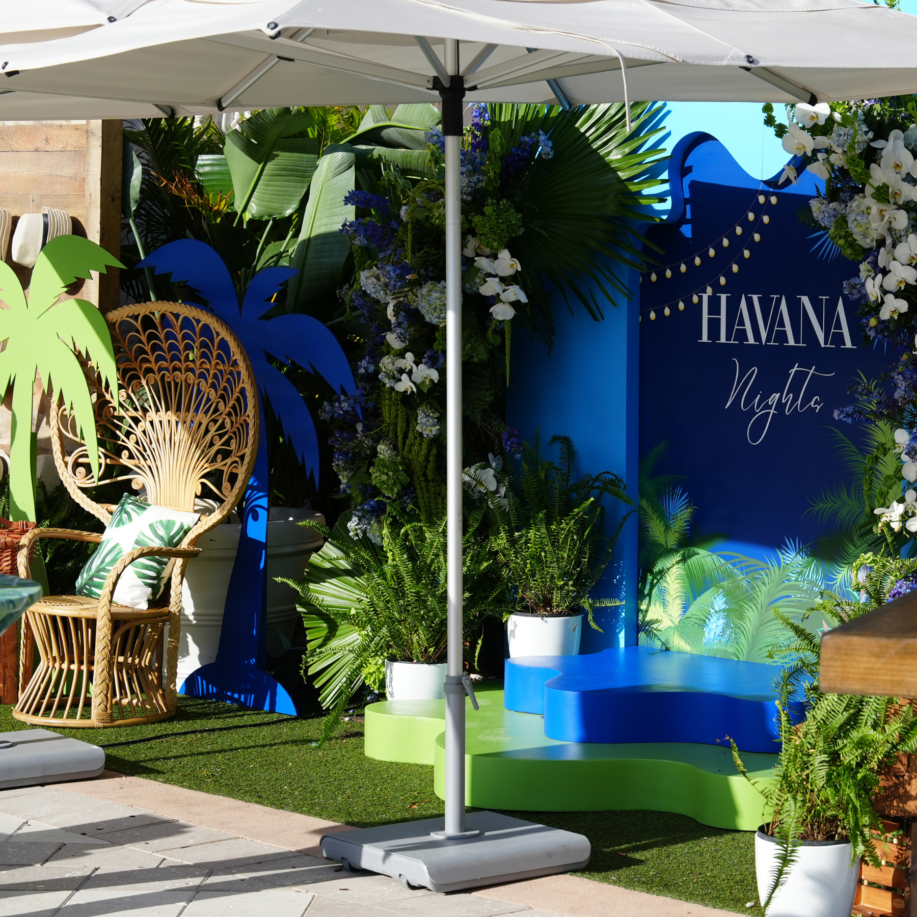 Outdoor tropical-themed decoration with plants, a wicker chair with a leaf-patterned pillow, and a blue and green platform under a large white umbrella, with a sign reading 'Havana Nights' surrounded by floral arrangements.