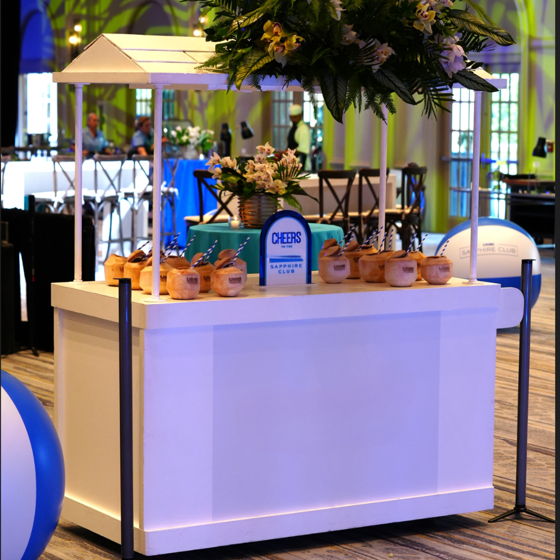 A white island bar with ten coconuts and striped straws, decorated with a sign that reads "Cheers to the Sapphire Club," and a large flower arrangement on top, situated in a brightly lit event space.