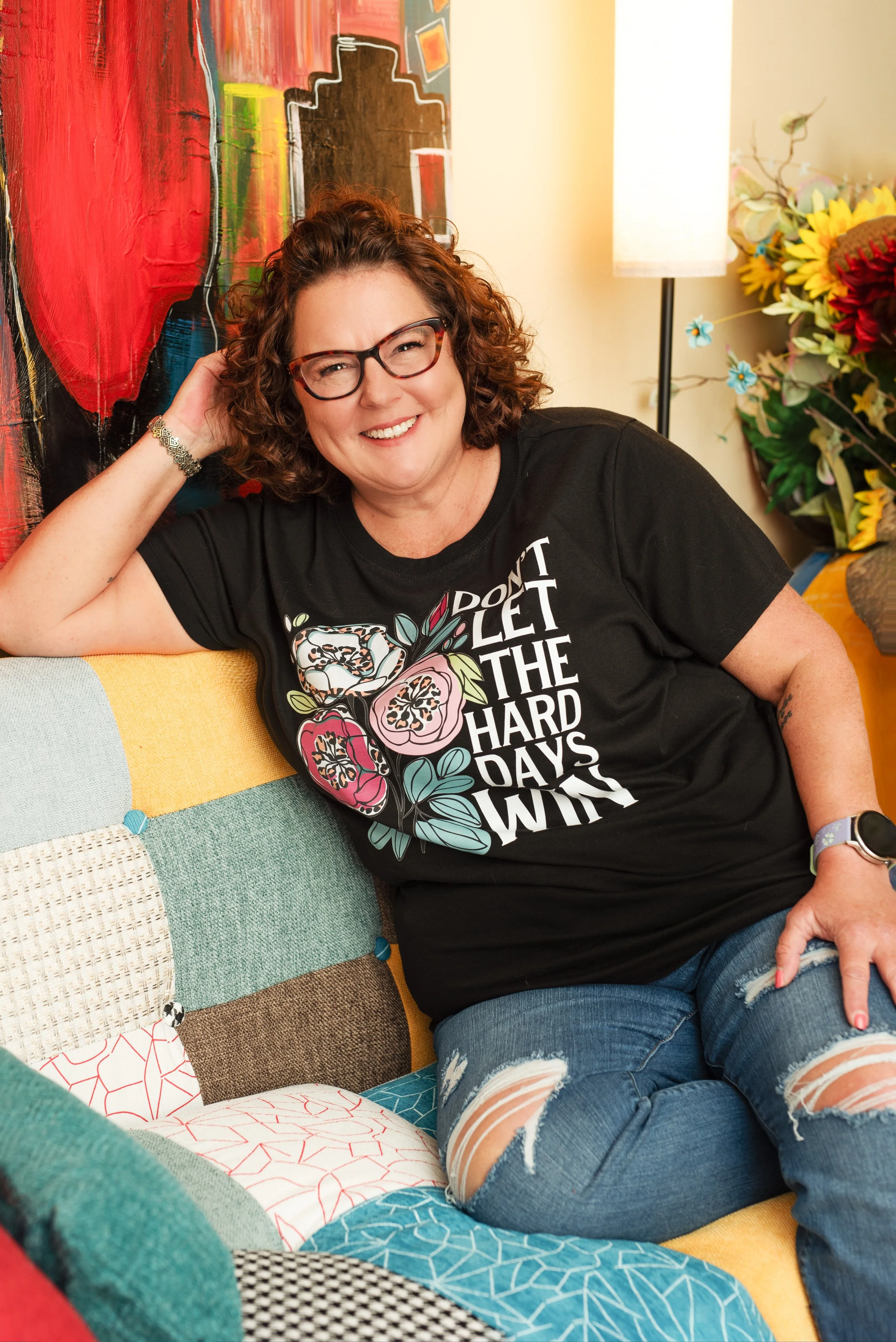 A smiling woman with curly hair and glasses, wearing a black T-shirt with floral design and text, sitting on a colorful patchwork couch, with a background of wall art, a lamp, and a bouquet of sunflowers and other flowers.