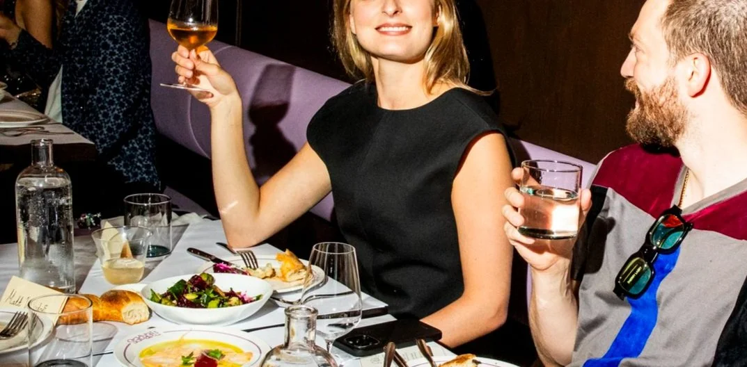 Two people enjoying a dinner party, clinking glasses with drinks, seated at a nicely set table with salad, bread, and a glass of wine.