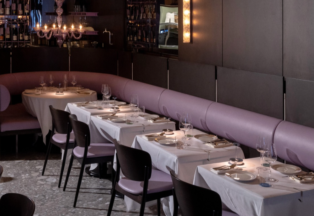 Restaurant interior with a long table set with white tablecloths, glassware, and silverware, pink cushioned booth seating along the wall, and a chandelier hanging from the ceiling.