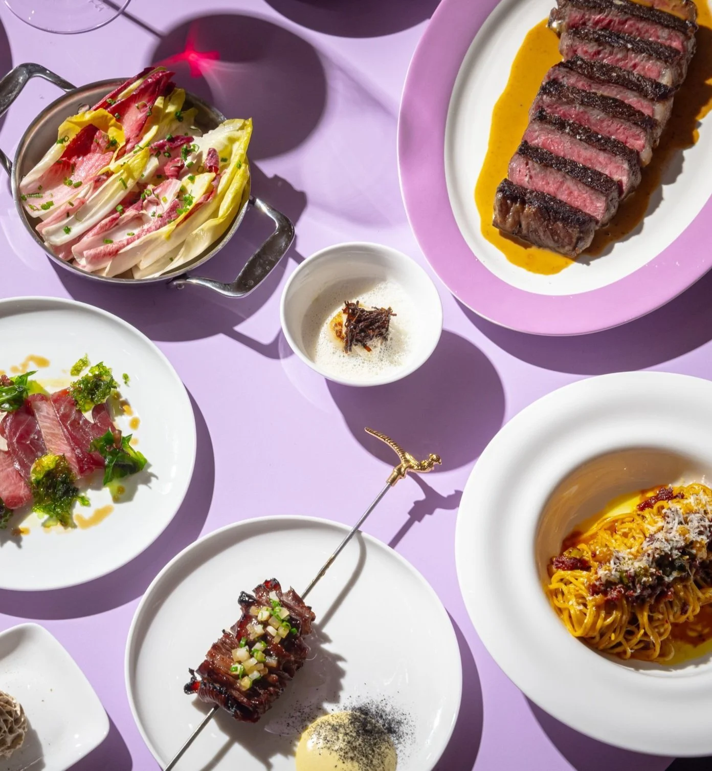 Assorted dishes including a grilled steak with sauce, raw meat with greens, a skewer of grilled meat with chopped onions, a vegetable salad, a small bowl with foam and garnish, a plate of pasta with sauce, and another plate with sliced meat greens, a