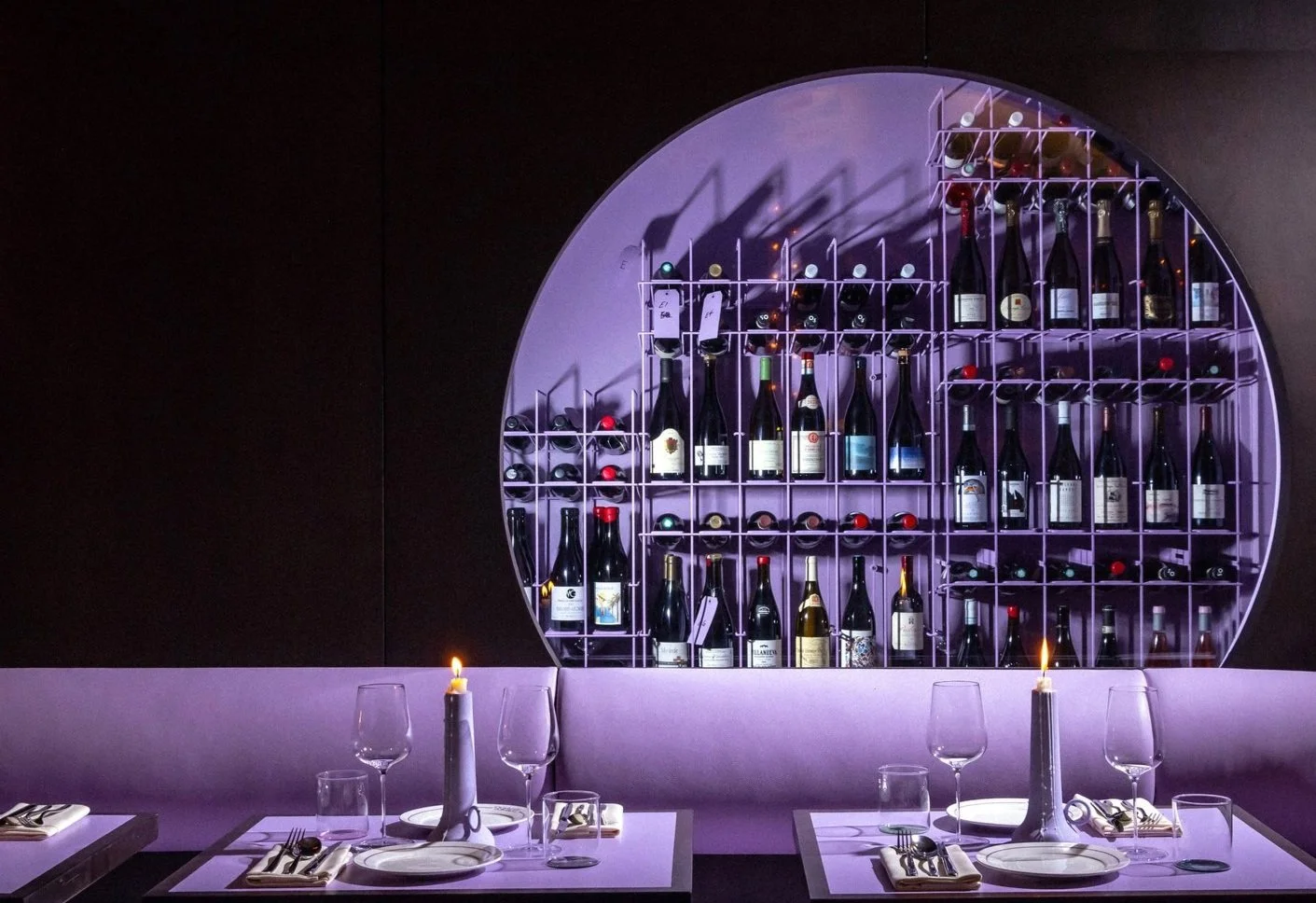 A modern restaurant interior with a purple-lit circular wine display filled with wine bottles, surrounded by tables set for dining with candles, glassware, plates, and utensils.