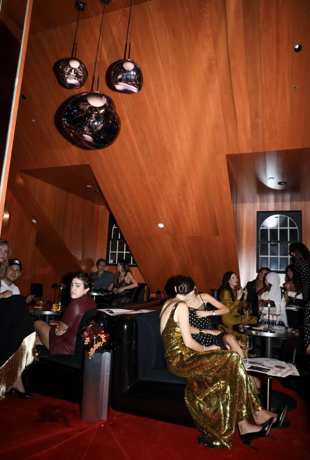 People sitting on sofas and chairs in a wood-paneled room, socializing and enjoying drinks. The room has large black pendant lights hanging from the ceiling and arched windows in the background. Some women are dressed in elegant party dresses, includ