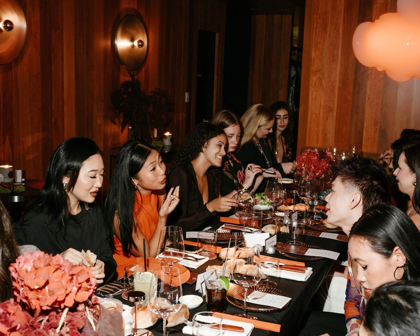 People sitting at a long dinner table in the dimly lit PearlBox in SoHo, enjoying a social meal with drinks and flowers.