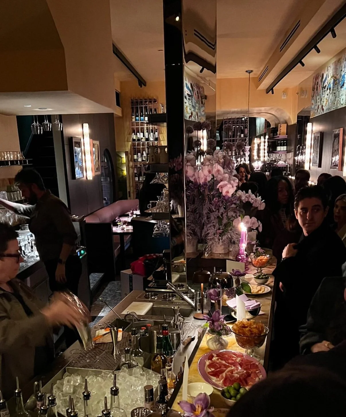 A lively restaurant bar with inside decor, a bartender preparing drinks, and patrons enjoying their time, with a large arrangement of pink flowers and various food dishes on the counter.