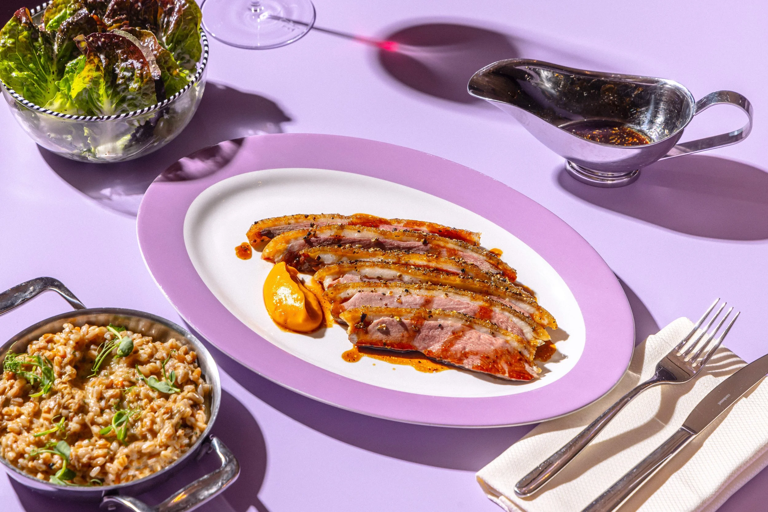 Sliced seared duck breast with sauce on a white and purple plate, side of risotto with herbs in a small cast iron skillet, green salad in a glass bowl, gravy boat with sauce, and a glass of red wine on a lavender tablecloth with silverware and napkin