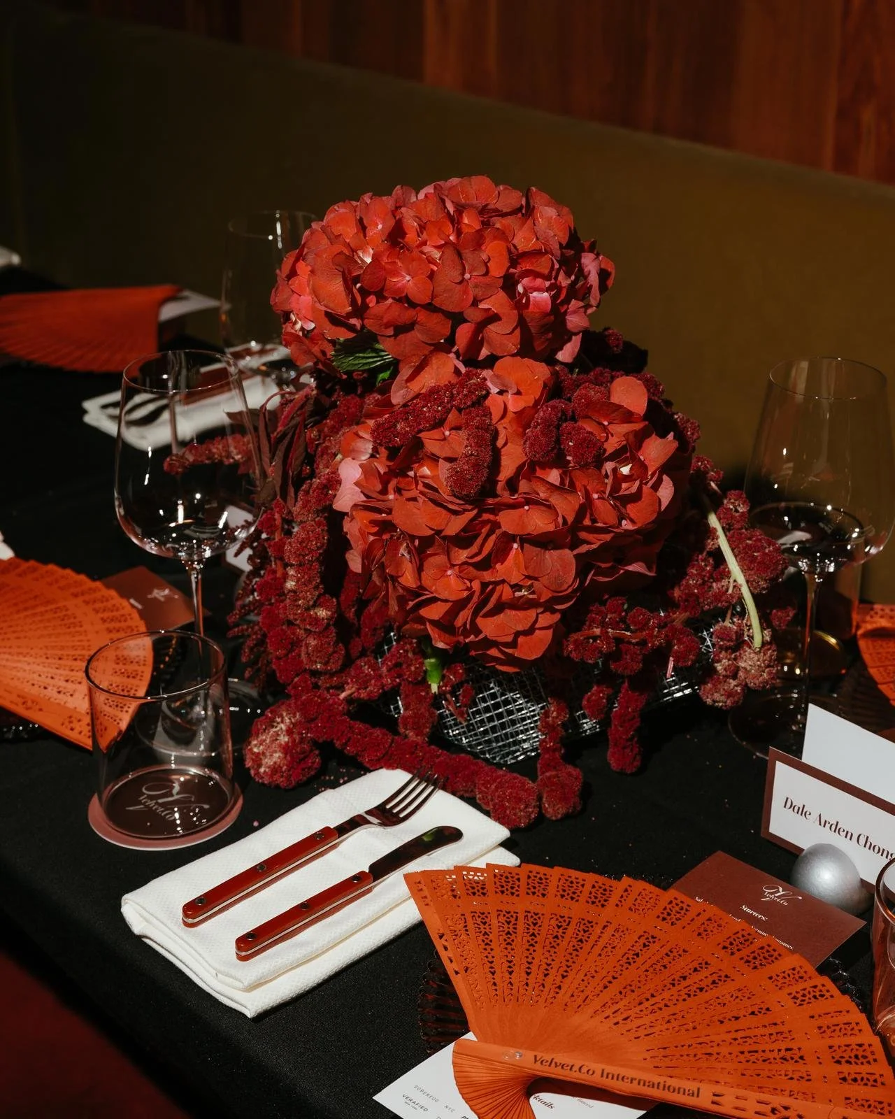 Elegant dining table with a floral centerpiece of red and orange flowers, surrounded by wine glasses, cutlery, and orange decorative fans.