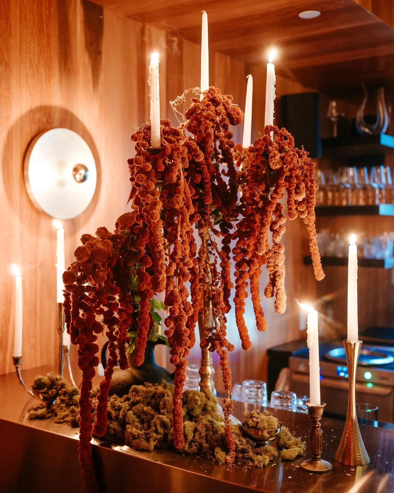 A floral centerpiece inside a private event space in NYC, with orange and red hanging amaranthus and white candles on a wooden table.